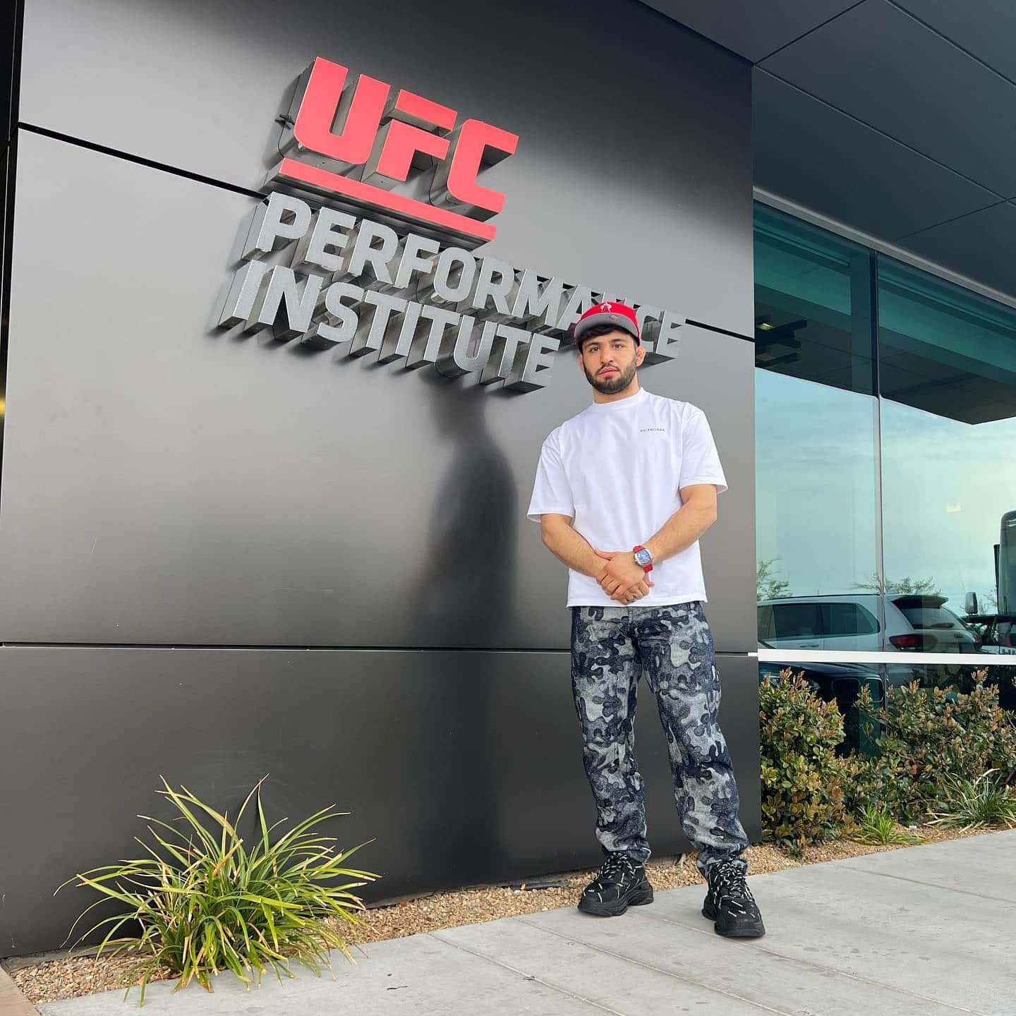 MMA 桌布 壁紙 — 一名身穿迷彩褲和紅色帽子的男子自信地站在UFC Performance Institute的現代玻璃和鋼結構外牆前。