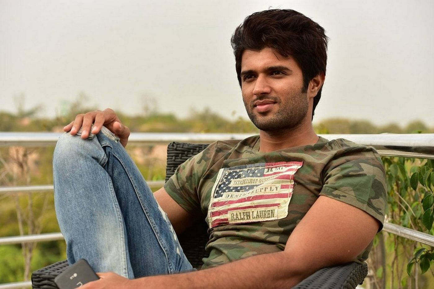 vijay devarakonda 4K 桌布 壁紙 — 一名身穿迷彩襯衫和牛仔褲的年輕男子放鬆地坐在陽台椅子上，手持電話，身後有綠植和欄杆。