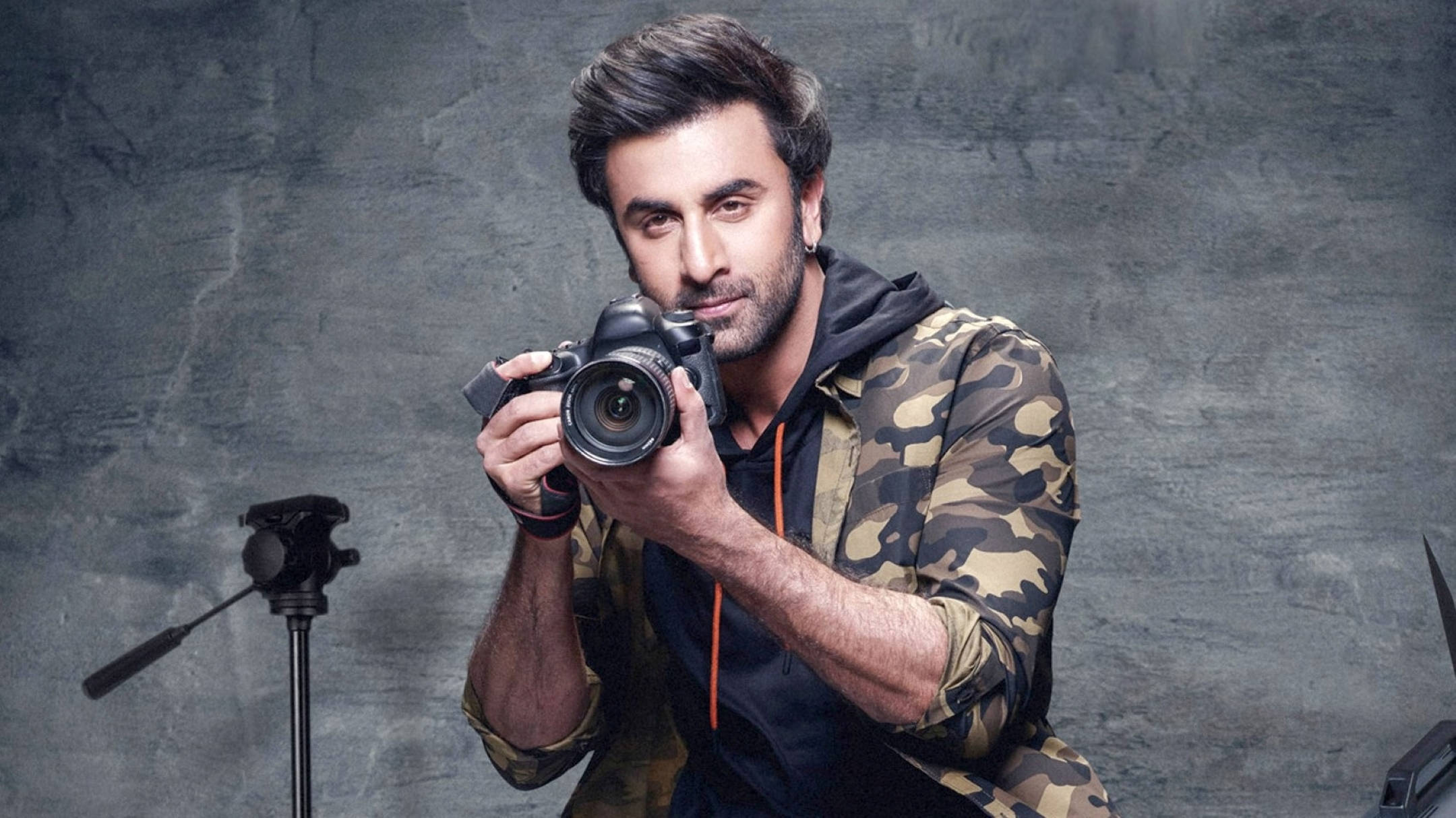 Ranbir Kapoor 桌布 壁紙 — 一名身穿迷彩連帽上衣的男子手持單眼相機，在帶有紋理的灰色攝影棚背景前直視觀眾。