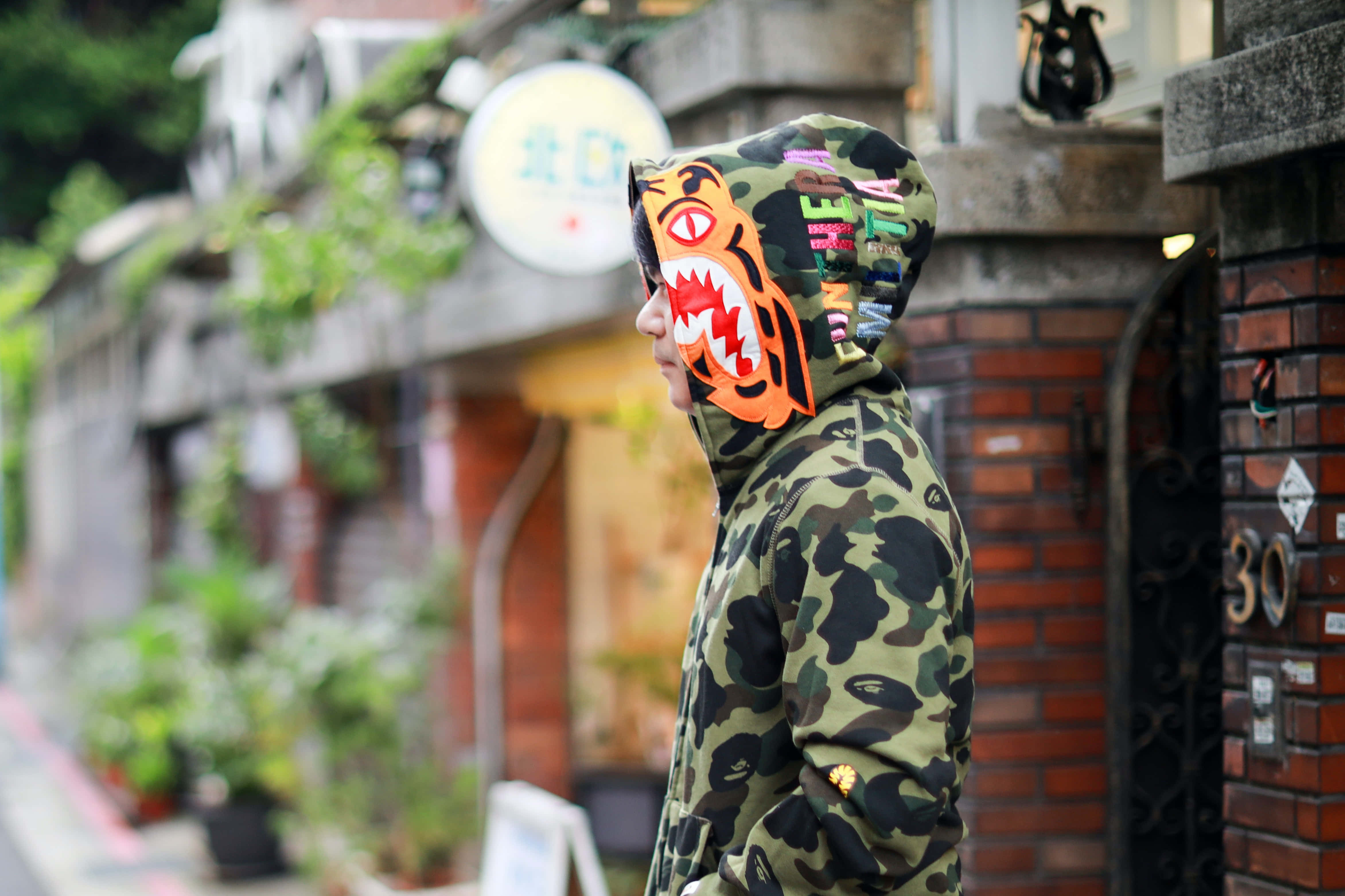 Bape 動態 桌布 壁紙 — 一名身穿迷彩連帽衫、戴著鮮橙色鯊魚面罩的人走在一條狹窄的磚砌街道上。
