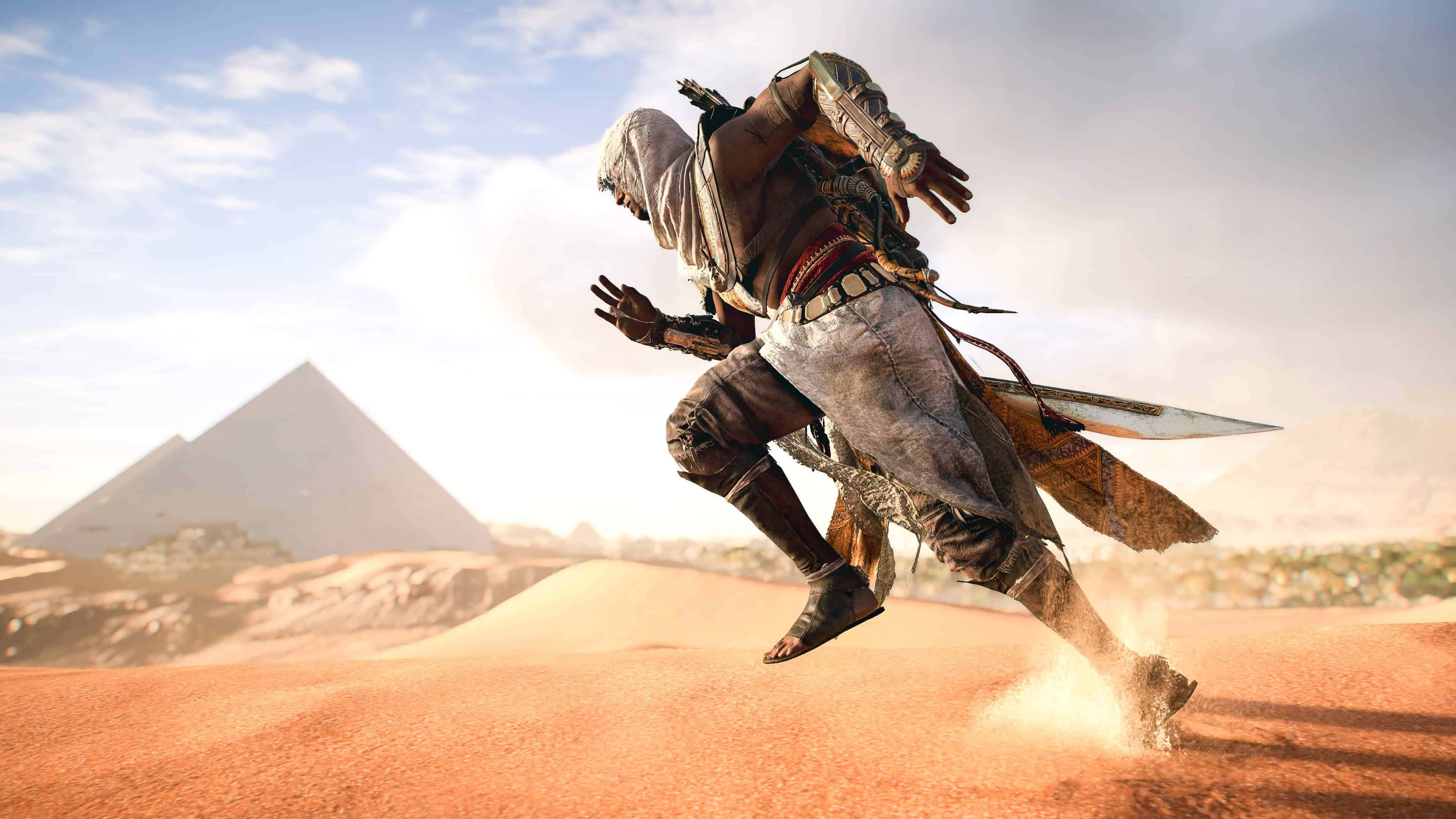 Bayek 桌布 壁紙 — 一名身穿連帽長袍的刺客在陽光明媚的沙漠中躍起，遠處朦朧中金字塔聳立。