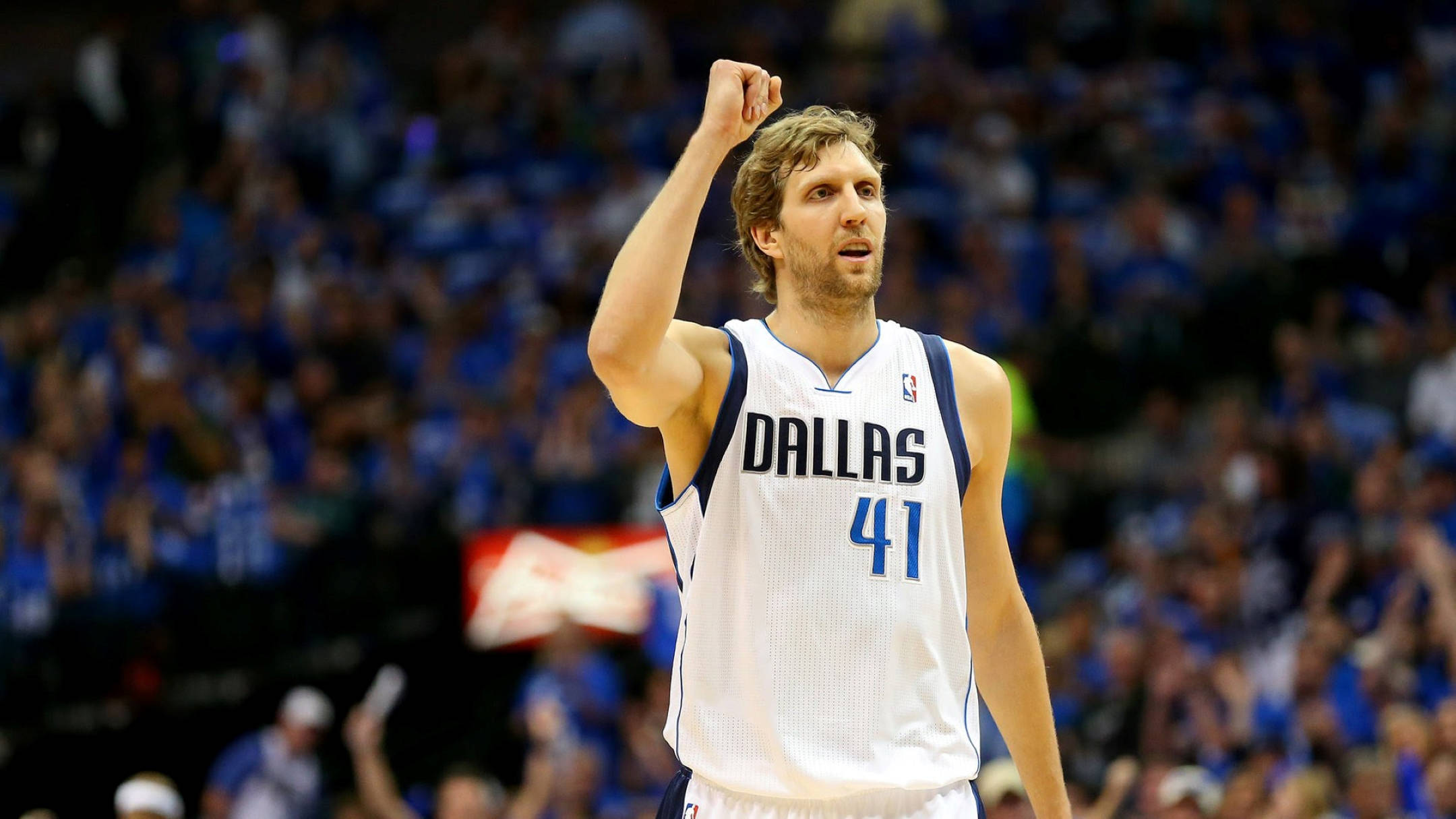 Dirk Nowitzki 桌布 壁紙 — 一名身穿達拉斯球衣的籃球運動員在場館燈光下，於歡呼的人群中高舉拳頭慶祝勝利。