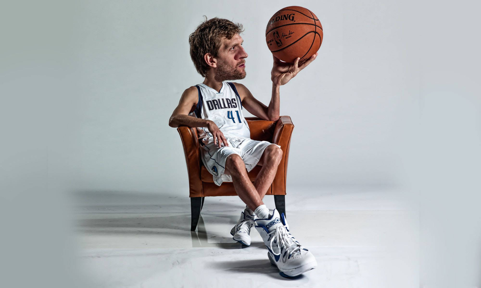 Dirk Nowitzki 桌布 壁紙 — 一名身穿達拉斯球衣的籃球運動員放鬆地坐在棕色皮椅上，手持籃球，背景是純白色攝影棚。