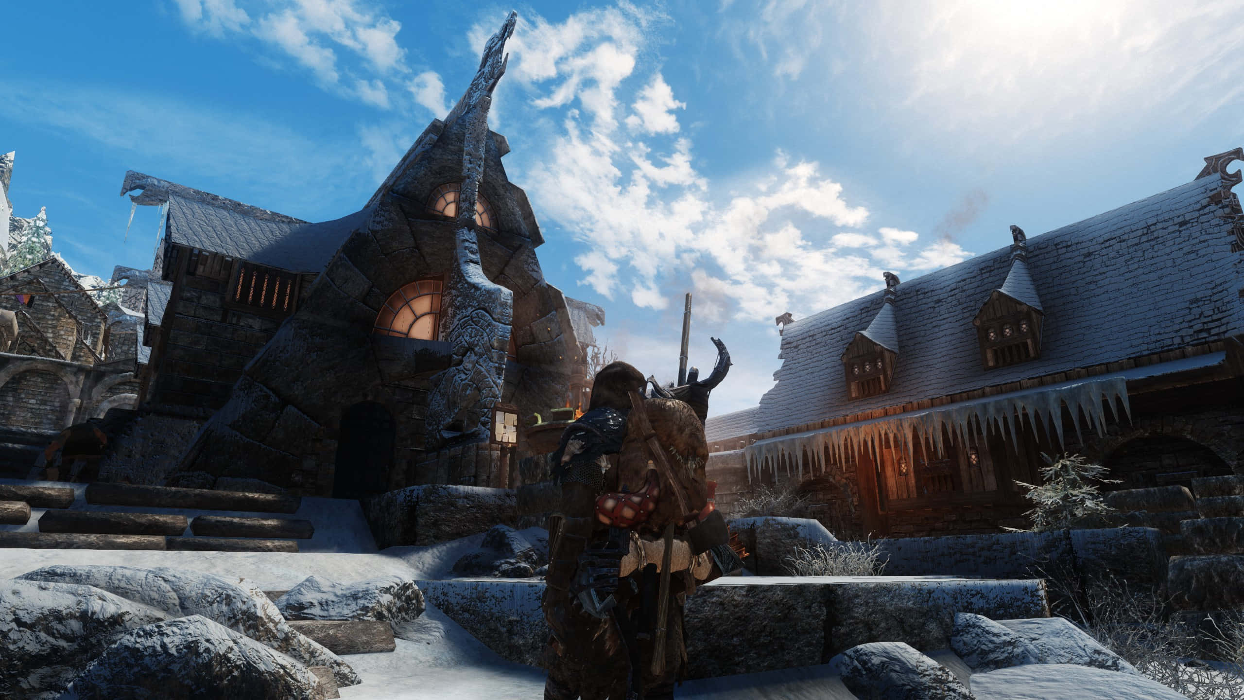Skyrim Windhelm 桌布 壁紙 — 一名身穿重甲的戰士站在白雪覆蓋的中世紀村莊中，村莊有石頭建築和冰柱，背景是明亮的藍天。