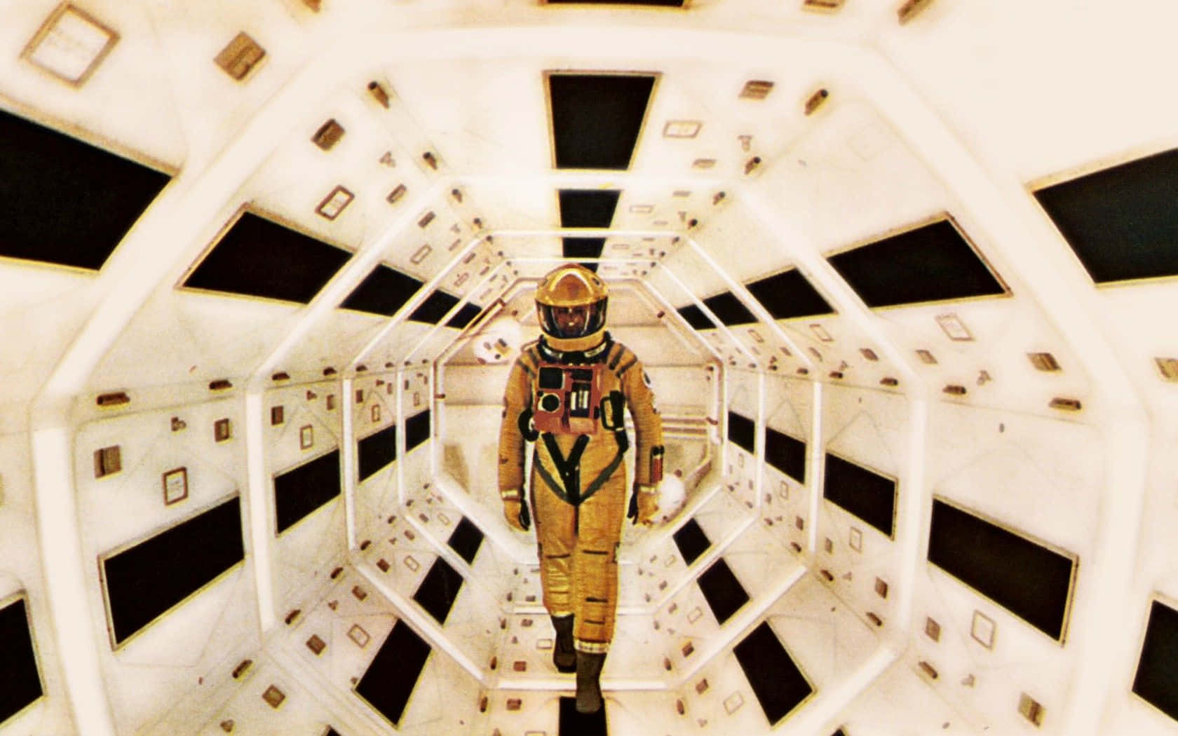 2001 A Space Odyssey 桌布 壁紙 — 一名身穿金色太空衣的太空人，沿著一條狹窄的白色六角形太空船走廊行走，走廊上有黑色面板和小型方形裝置。