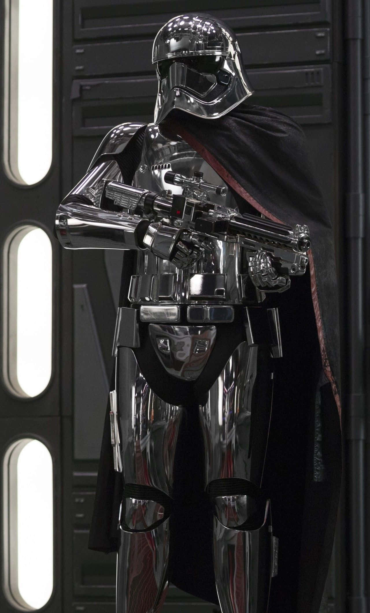 Captain Phasma 桌布 壁紙 — 一名身穿鉻合金裝甲的風暴兵披著黑色斗篷，站在光線昏暗的太空船走廊中，手持爆能槍。