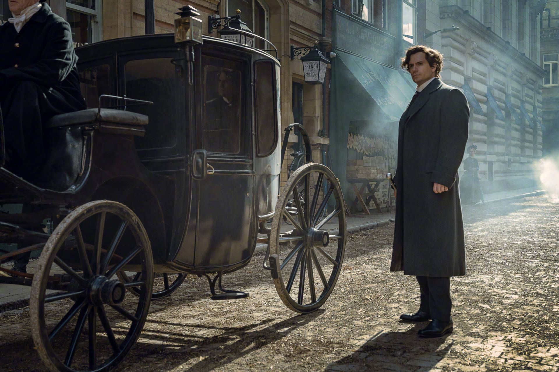 Henry Cavill 桌布 壁紙 — 一名身穿長外套的男子站在一輛復古馬車旁，身處一條塵土飛揚的鵝卵石街道上，沐浴在金色的晨光中。