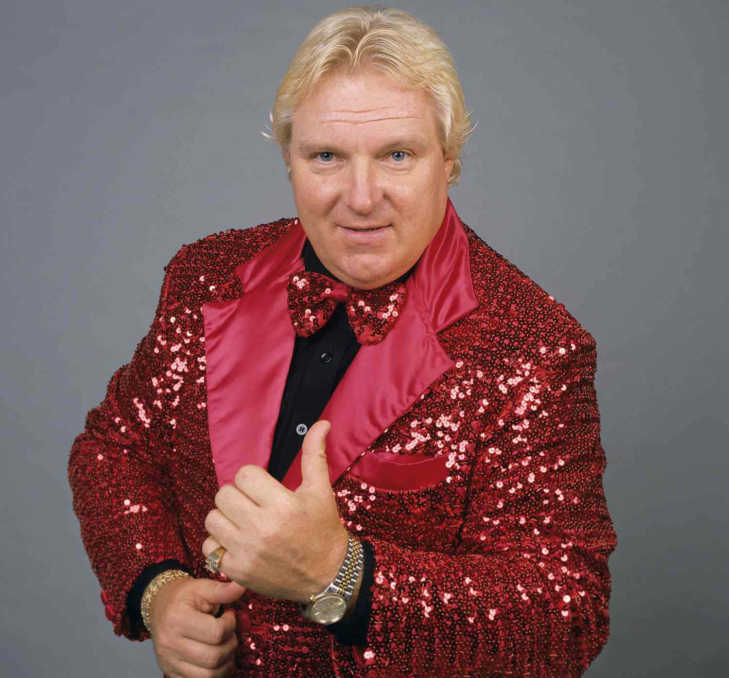 Bobby Heenan 桌布 壁紙 — 一名身穿閃亮紅色亮片外套和相配領結的男子，在純灰色背景前自信地豎起大拇指。