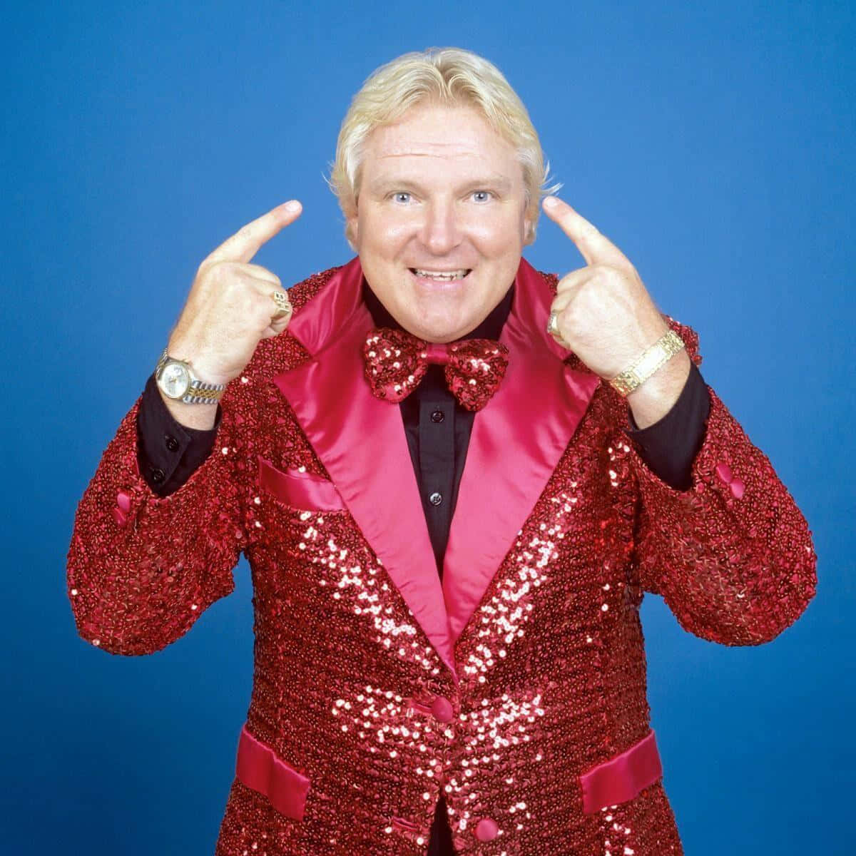 Bobby Heenan 桌布 壁紙 — 一名身穿閃亮紅色亮片西裝、搭配相襯領結的男子，在純藍色背景前指著自己的頭。