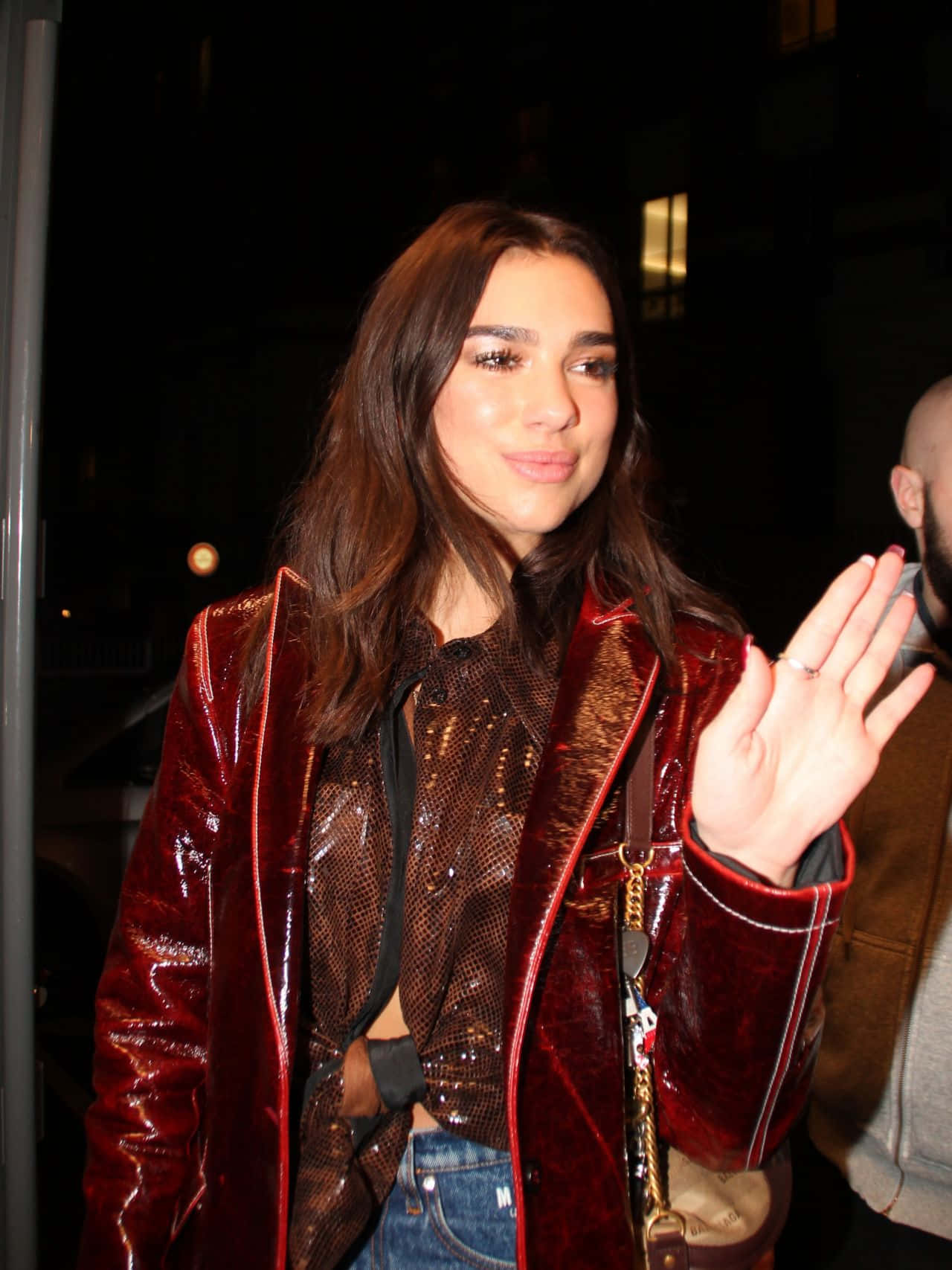 Dua Lipa 桌布 壁紙 — 一名身穿閃亮紅色外套的女性在夜間城市燈光下向鏡頭揮手。