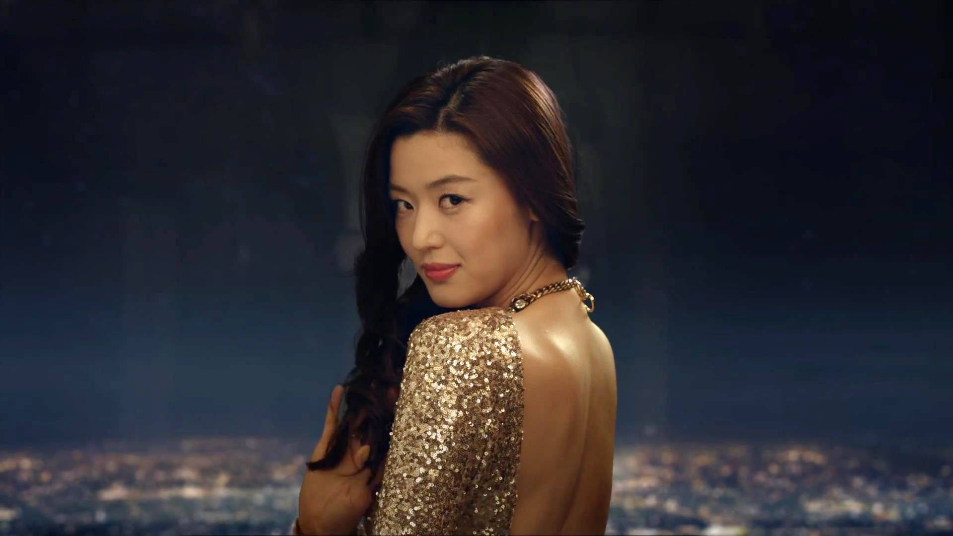 Jun Ji Hyun 桌布 壁紙 — 一名身穿閃亮金色連衣裙的女子在夜晚模糊的城市天際線背景下轉頭回眸。