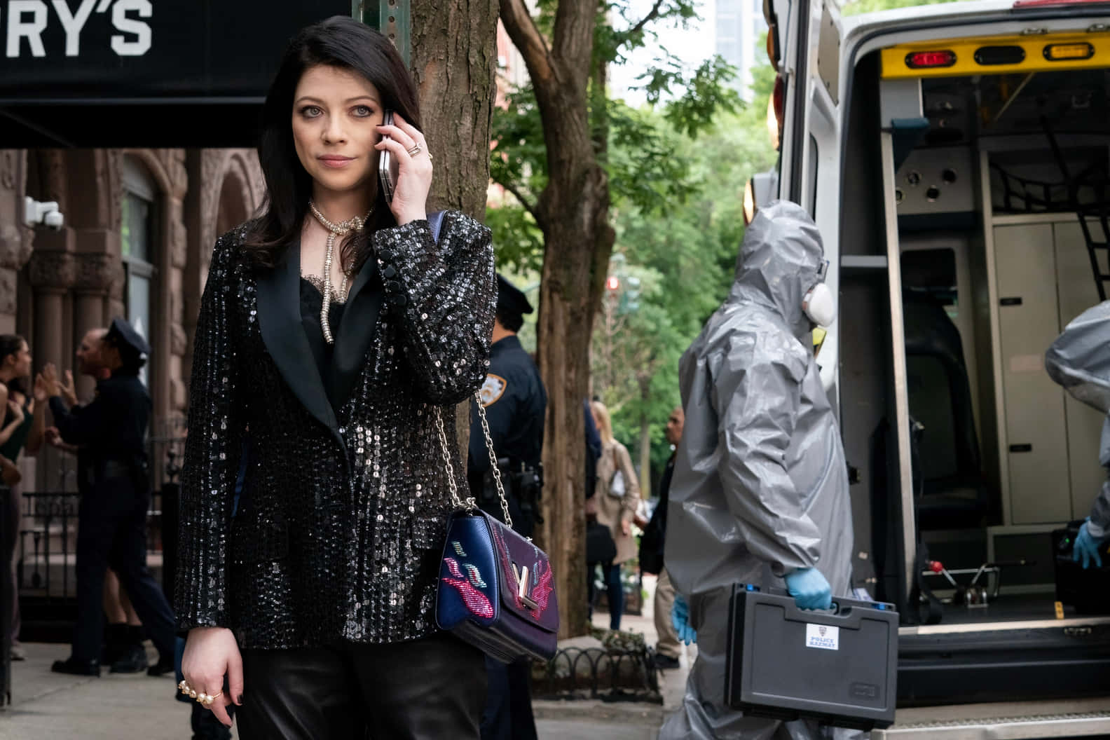 michelle trachtenberg 桌布 壁紙 — 一名身穿閃亮黑色西裝外套的女子正在講電話，而穿著防護服的鑑識小組在救護車旁的城市街道上工作。