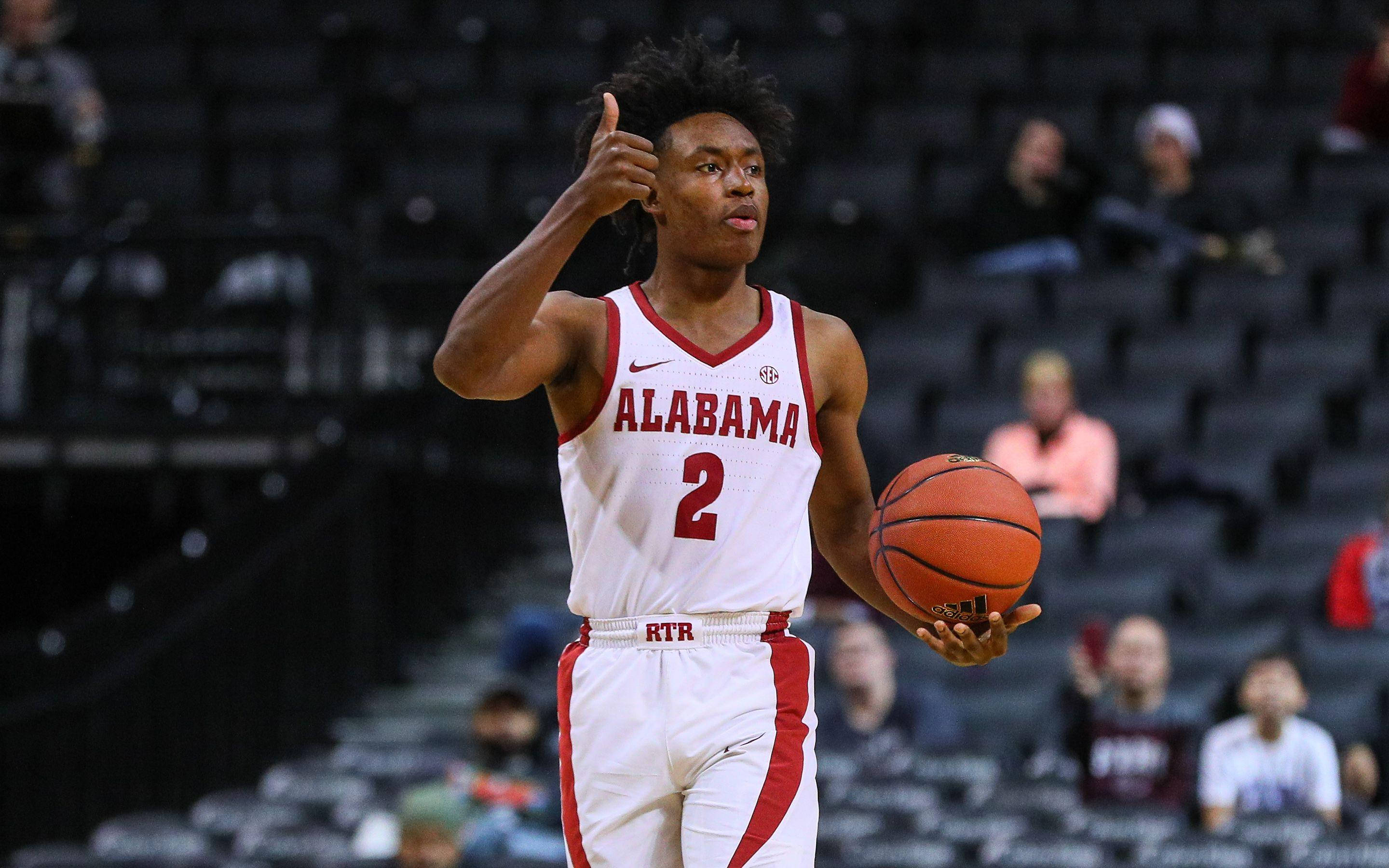 Collin Sexton 桌布 壁紙 — 一名身穿阿拉巴馬球衣的籃球運動員在觀眾稀疏的球場上拿著球並豎起大拇指。