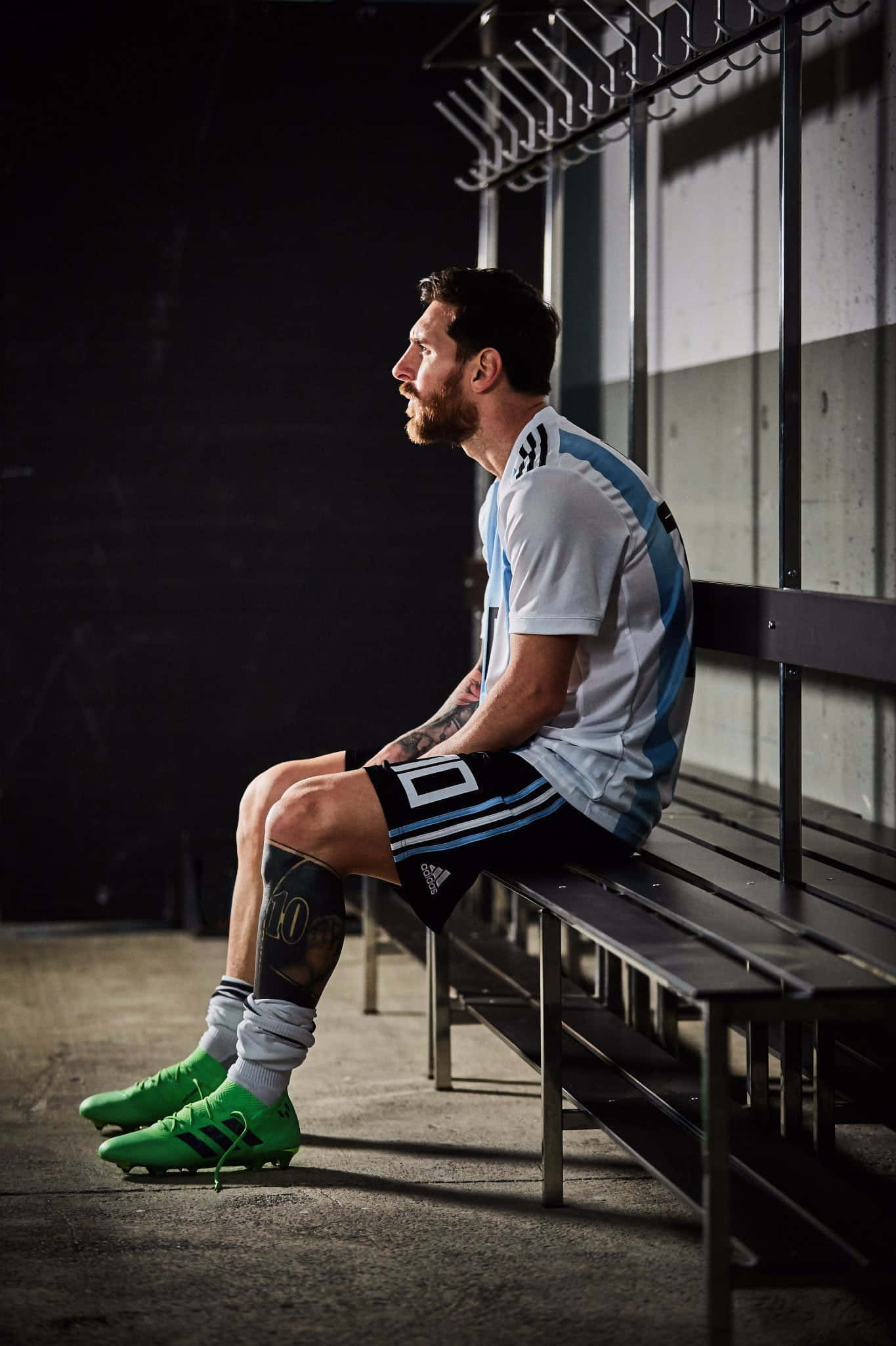 Messi 2022 桌布 壁紙 — 一名身穿阿根廷藍白球衣的足球員，在燈光昏暗的更衣室裡，若有所思地坐在金屬長凳上，穿著亮綠色球鞋。