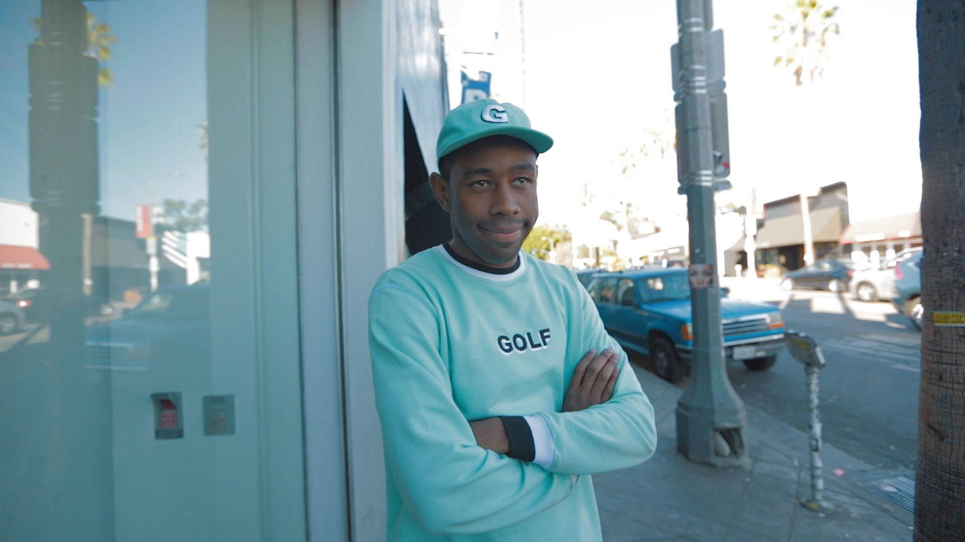 tyler 桌布 壁紙 — 一名身穿青綠色 Golf 運動衫和帽子的男子，在陽光明媚的城市人行道上靠著玻璃門，背景有棕櫚樹和藍色卡車。