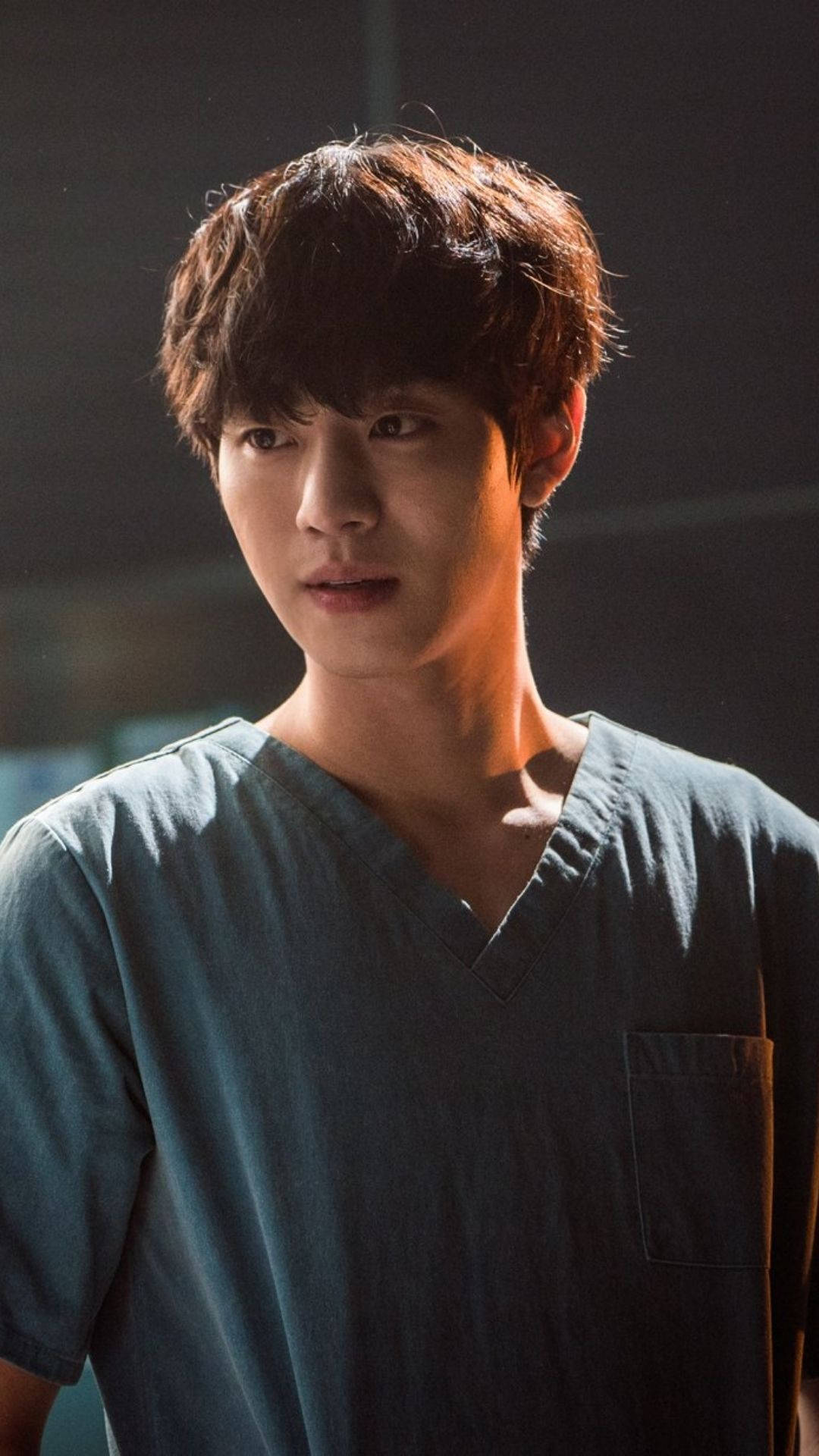 Ahn Hyo Seop 桌布 壁紙 — 一名身穿青綠色手術服的年輕男子站在昏暗燈光下，神情專注，棕色頭髮凌亂。