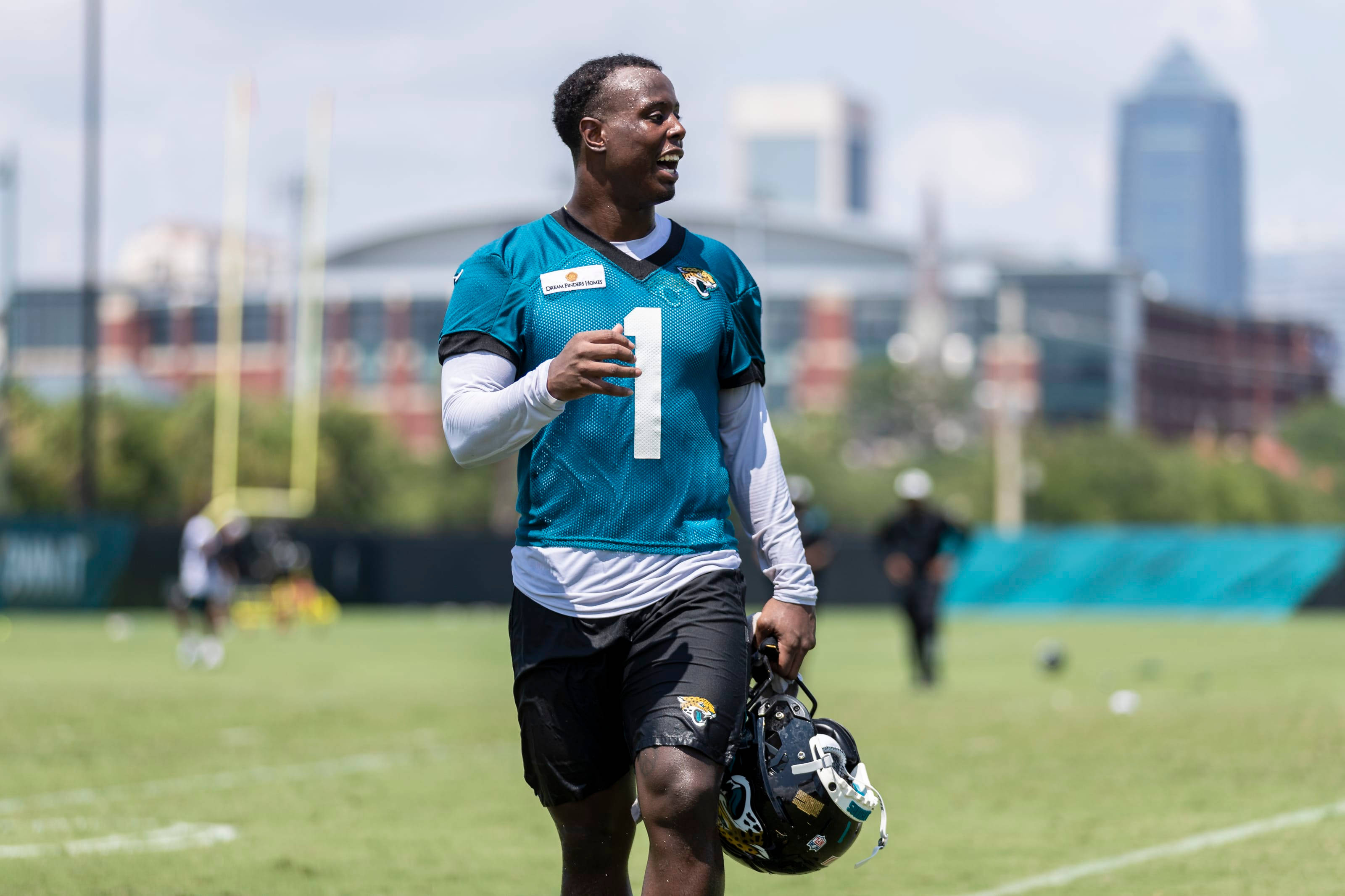 Travis Etienne 桌布 壁紙 — 一名身穿青綠色球衣一號的美式足球員在陽光明媚的球場上行走，手持頭盔，背景是城市天際線。