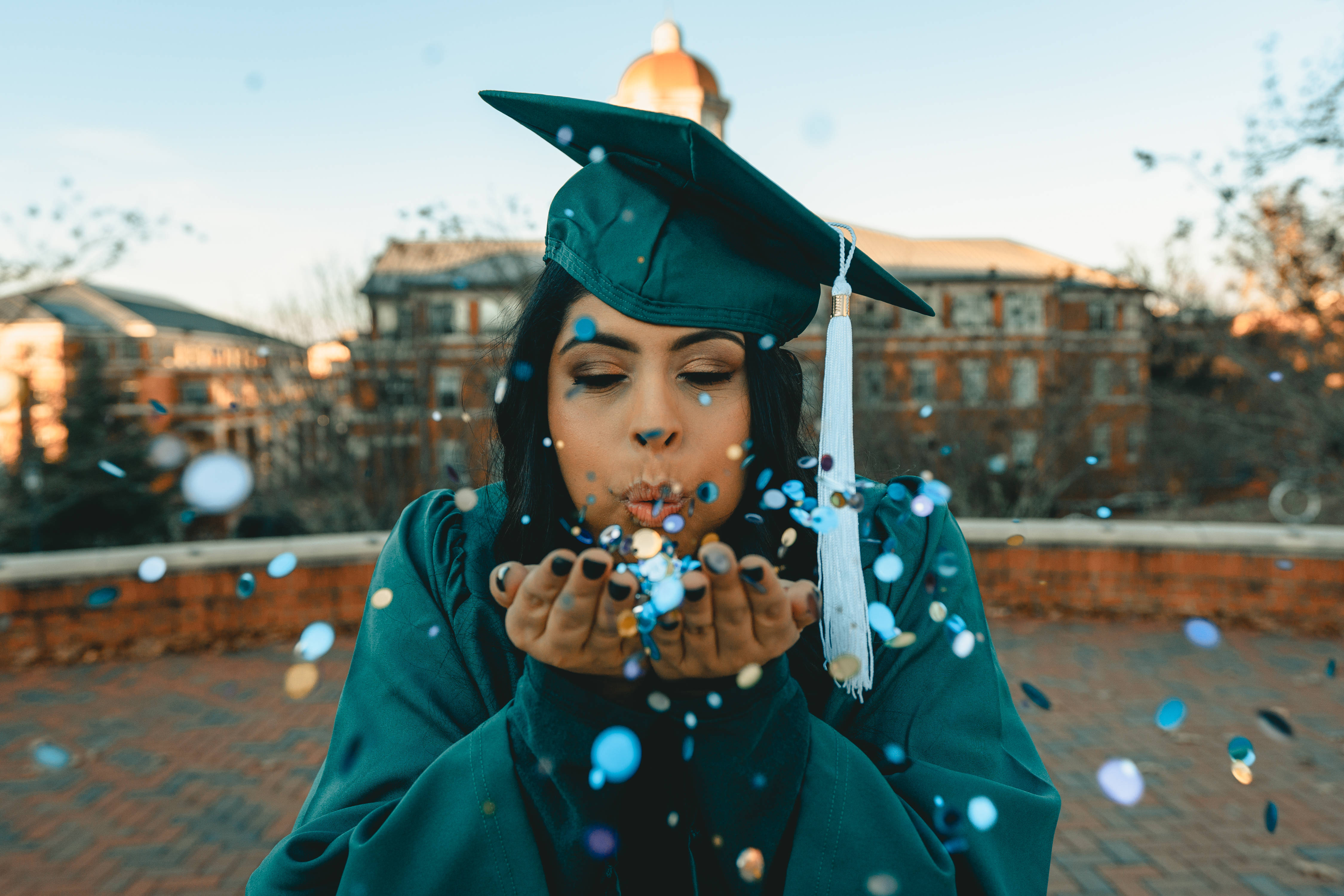 Success 桌布 壁紙 — 一名身穿青綠色畢業帽和袍的畢業生在黃金時段的大學校園背景下，將五彩紙屑吹向空中。
