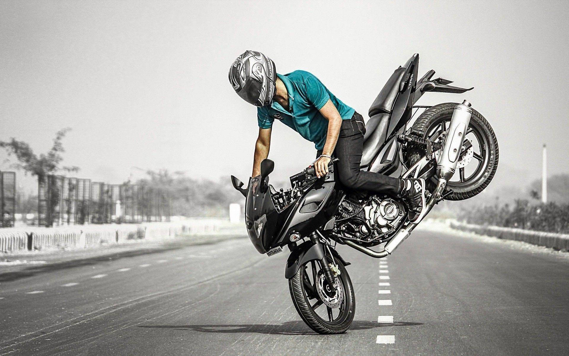 機車 桌布 壁紙 — 一名身穿青綠色襯衫的摩托車手，在陰霾天空下的荒涼道路上，騎著黑色跑車表演前輪離地。