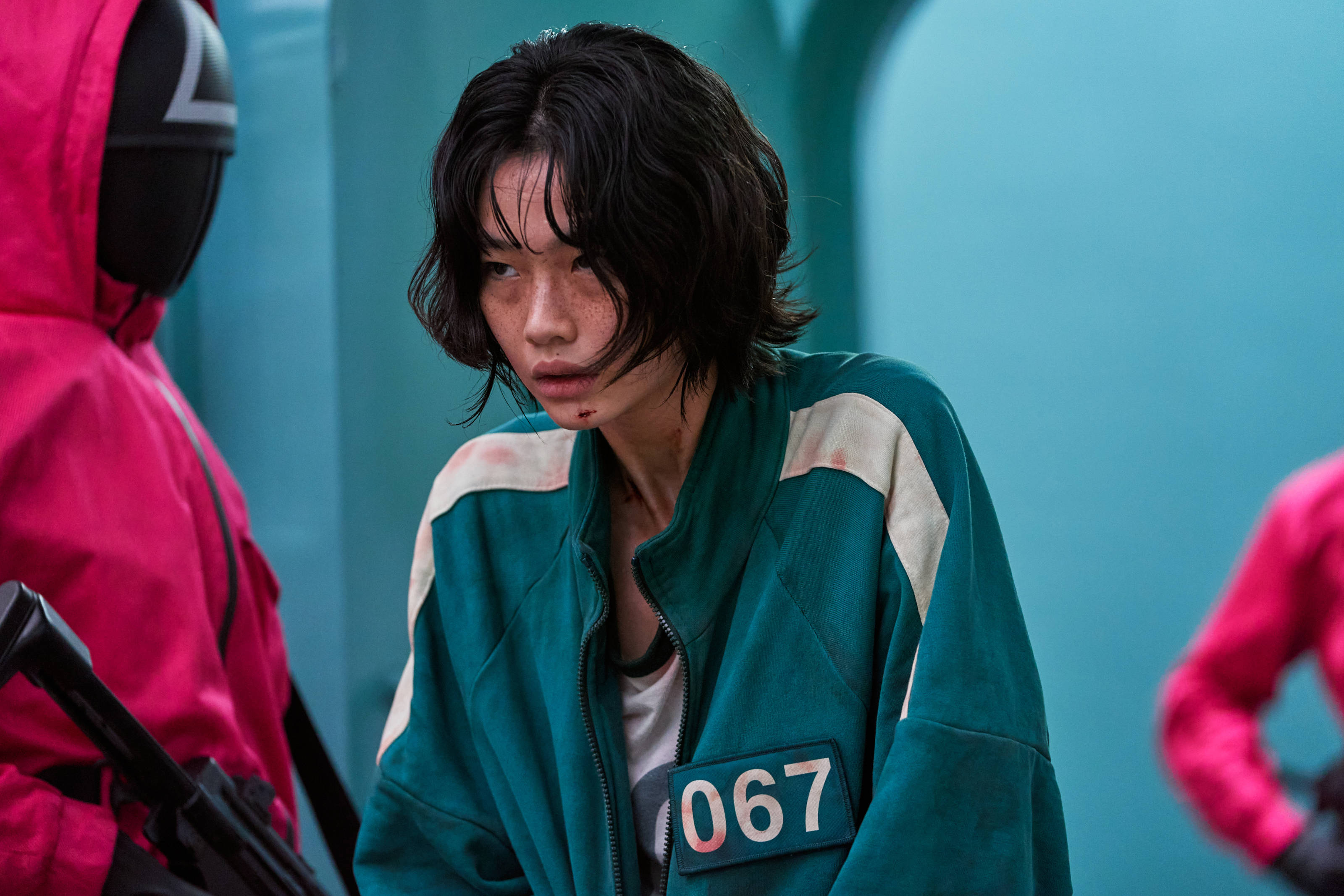 game 桌布 壁紙 — 一名身穿青綠色魷魚遊戲制服、編號067的苦惱玩家，正對著青綠色牆壁凝視，附近有身穿粉紅色衣服的對手。