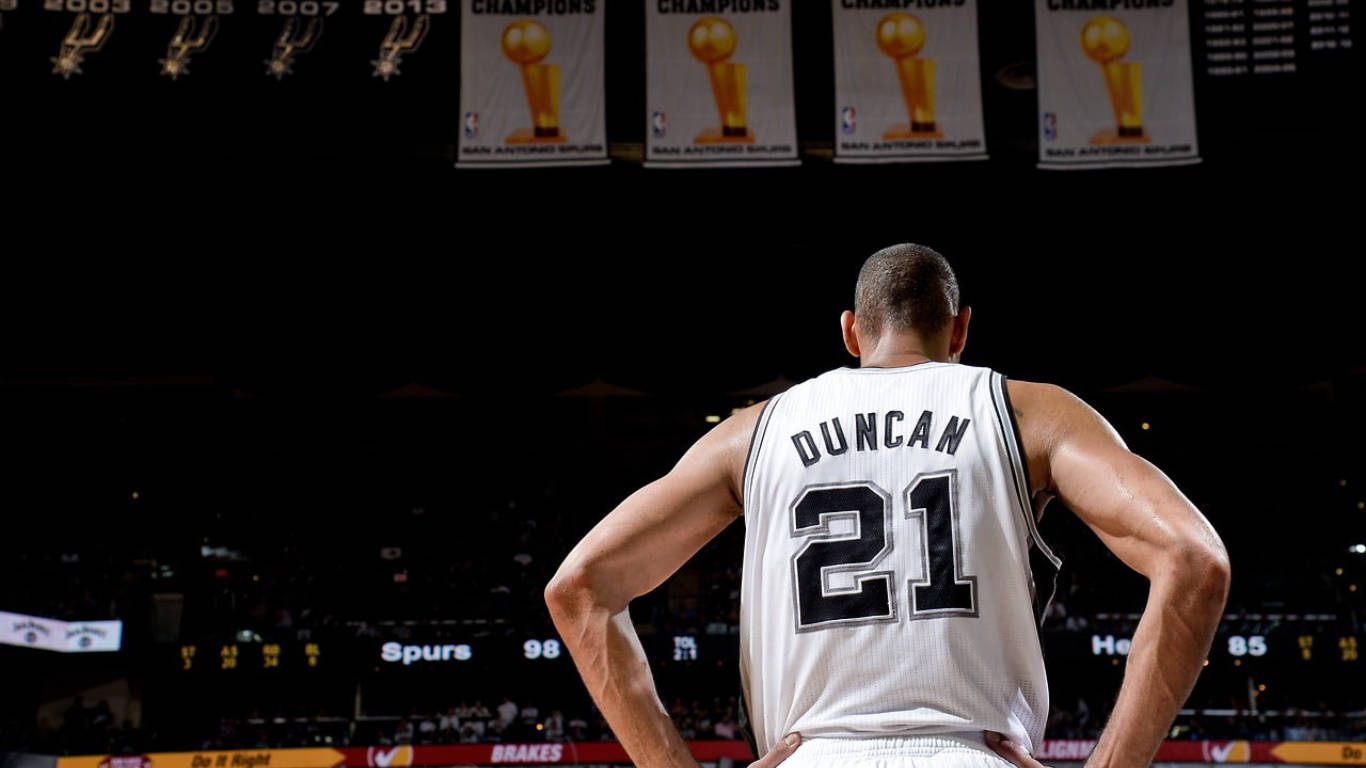 Spurs 桌布 壁紙 — 一名身穿馬刺球衣的籃球運動員在昏暗的球場中，雙手叉腰面向冠軍旗幟。