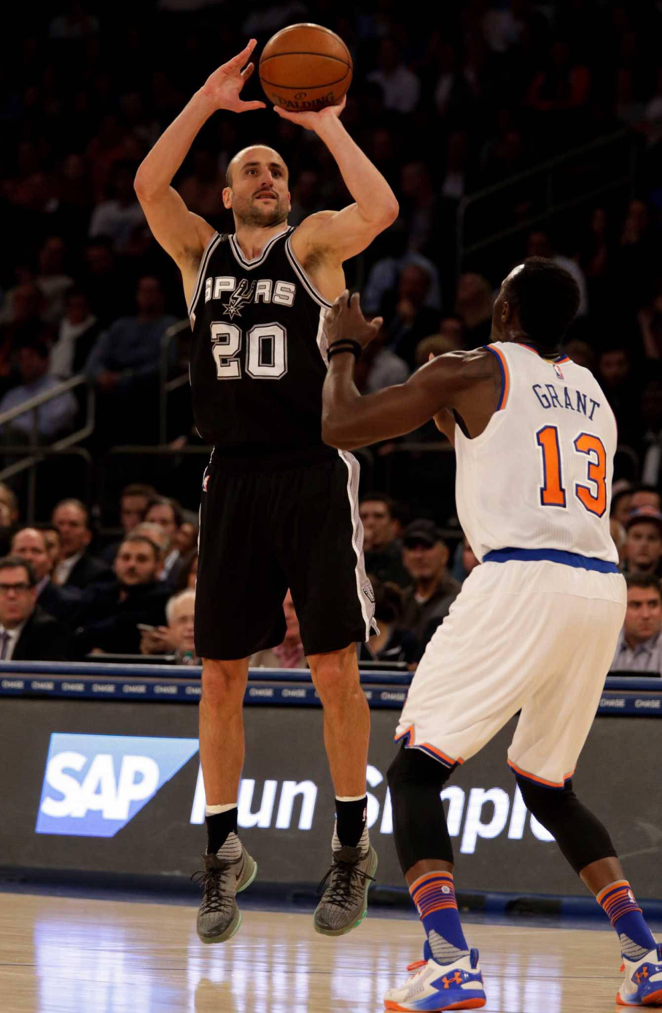 Manu 桌布 壁紙 — 一名身穿馬刺球衣的籃球運動員跳起投籃，而尼克防守球員在場館燈光下緊密防守他。