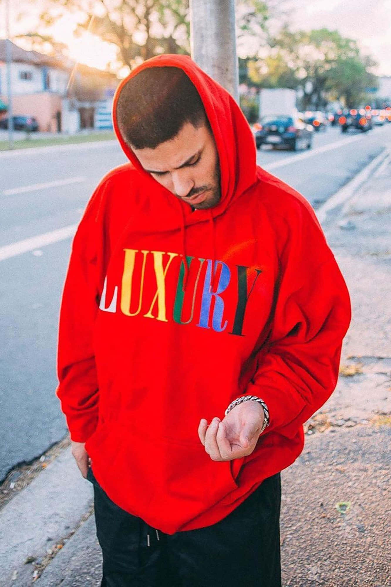 Red Hoodie 桌布 壁紙 — 一名身穿鮮紅色連帽衫、帶有彩虹字母的男子站在陽光明媚的城市人行道上，低頭看著自己的雙手。
