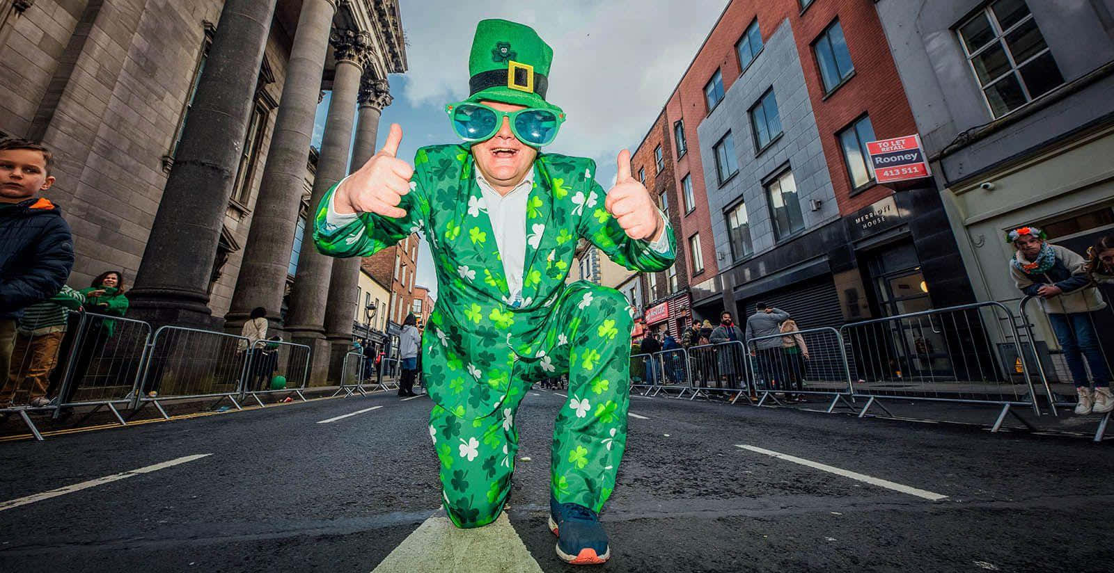 St Patrick's Day 桌布 壁紙 — 一名身穿鮮綠色三葉草西裝和帽子的男子在聖派翠克節遊行期間跑過城市街道，豎起兩個大拇指。