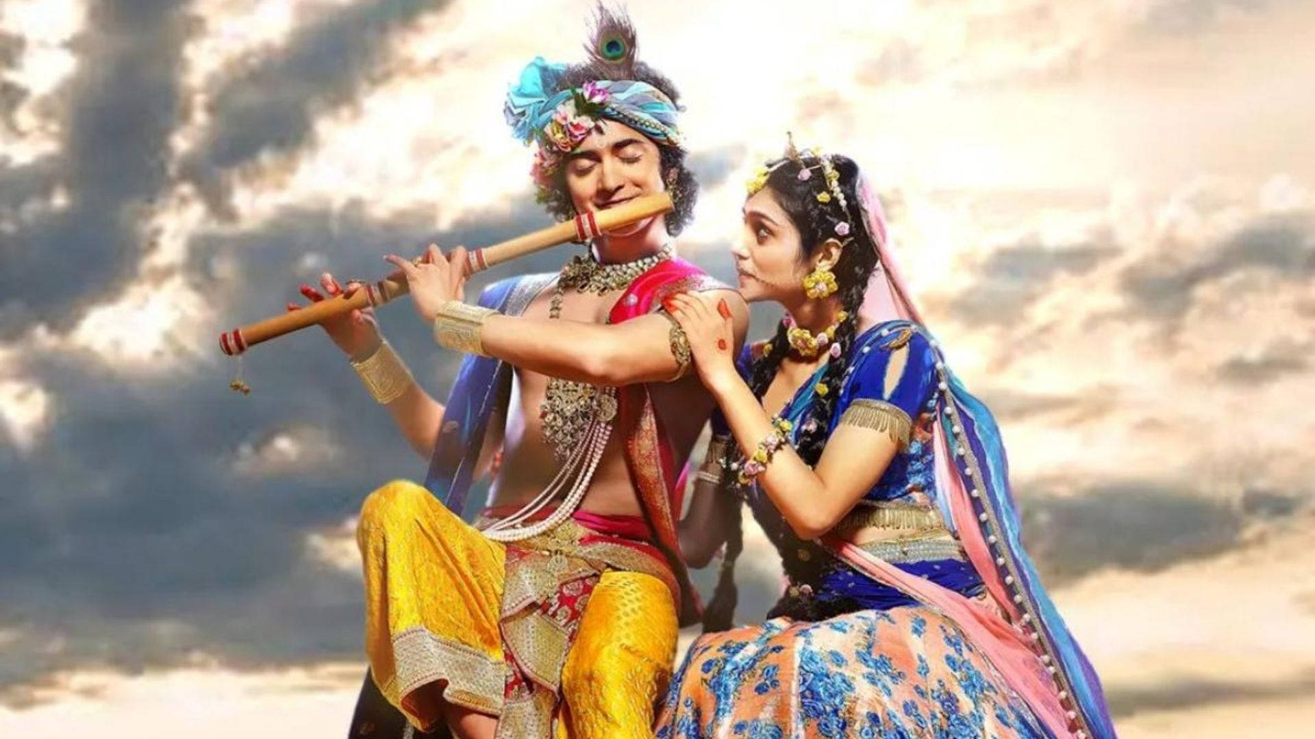 radha krishna serial 桌布 壁紙 — 一名身穿鮮豔傳統服飾的男子正在吹笛，而一名身穿華麗藍色和粉紅色連衣裙的女子在戲劇性的多雲天空下深情地靠在他身上。