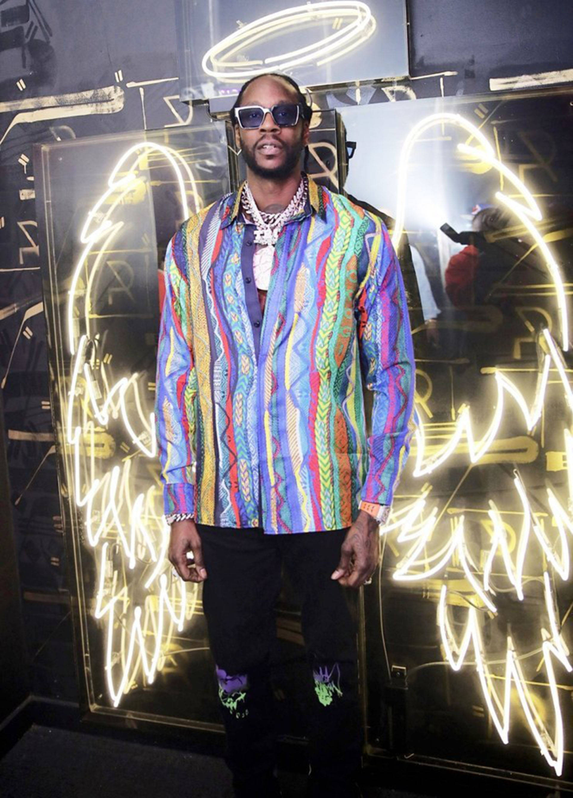 2 Chainz 桌布 壁紙 — 一名身穿鮮豔圖案襯衫和太陽眼鏡的男子自信地站在發光的霓虹藝術裝置前。