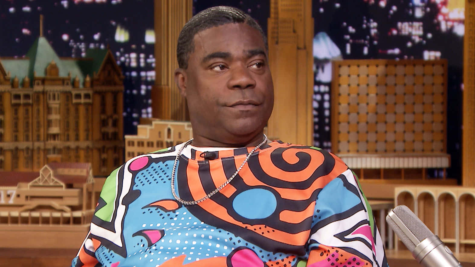 Tracy Morgan 桌布 壁紙 — 一名身穿鮮豔圖案襯衫的男子坐在脫口秀佈景中，背景是夜晚的城市天際線。
