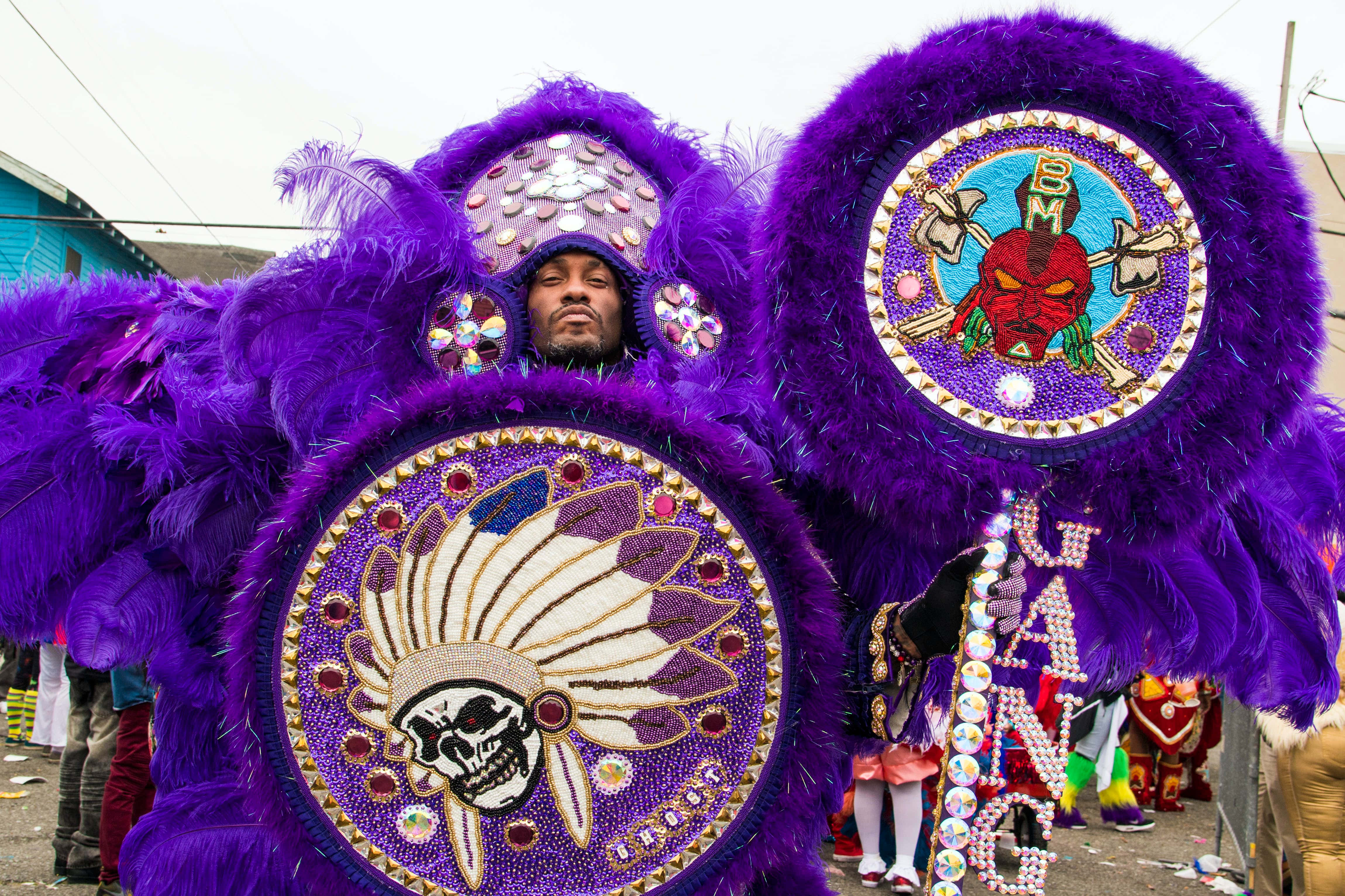 Mardi Gras 桌布 壁紙 — 一名身穿鮮豔紫色羽毛服飾、帶有珠飾頭骨和頭飾圖案的男人站在街頭遊行中。