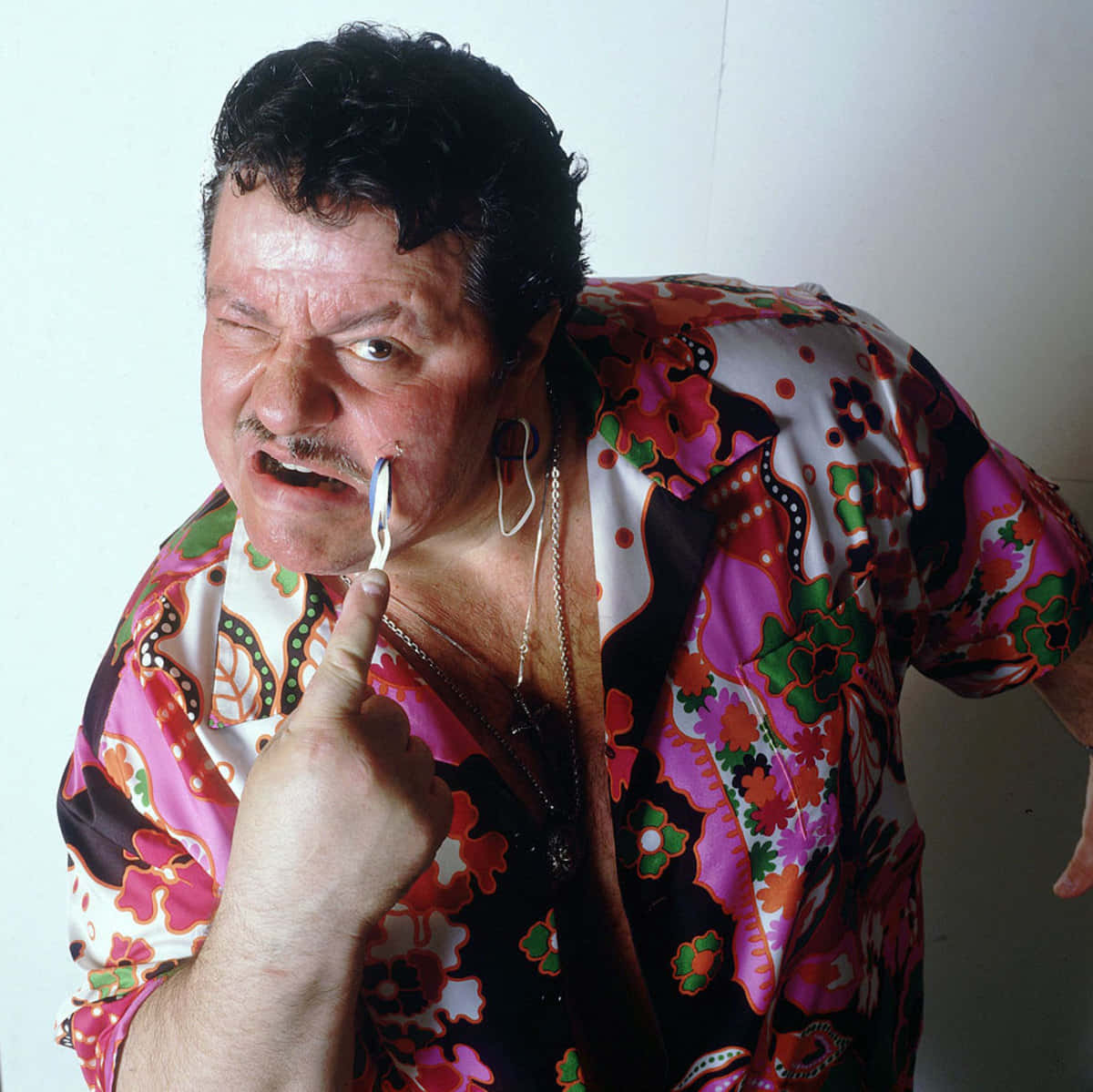 Lou Albano 桌布 壁紙 — 一名身穿鮮豔花卉襯衫的男子，在純白牆壁前眨眼並將剃刀刀片貼在臉頰上。