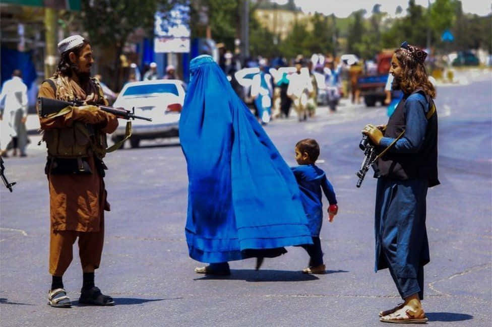 Taliban 桌布 壁紙 — 一名身穿鮮豔藍色布卡的女人帶著一個孩子行走，兩名武裝男子在陽光明媚的城市街道上站崗。