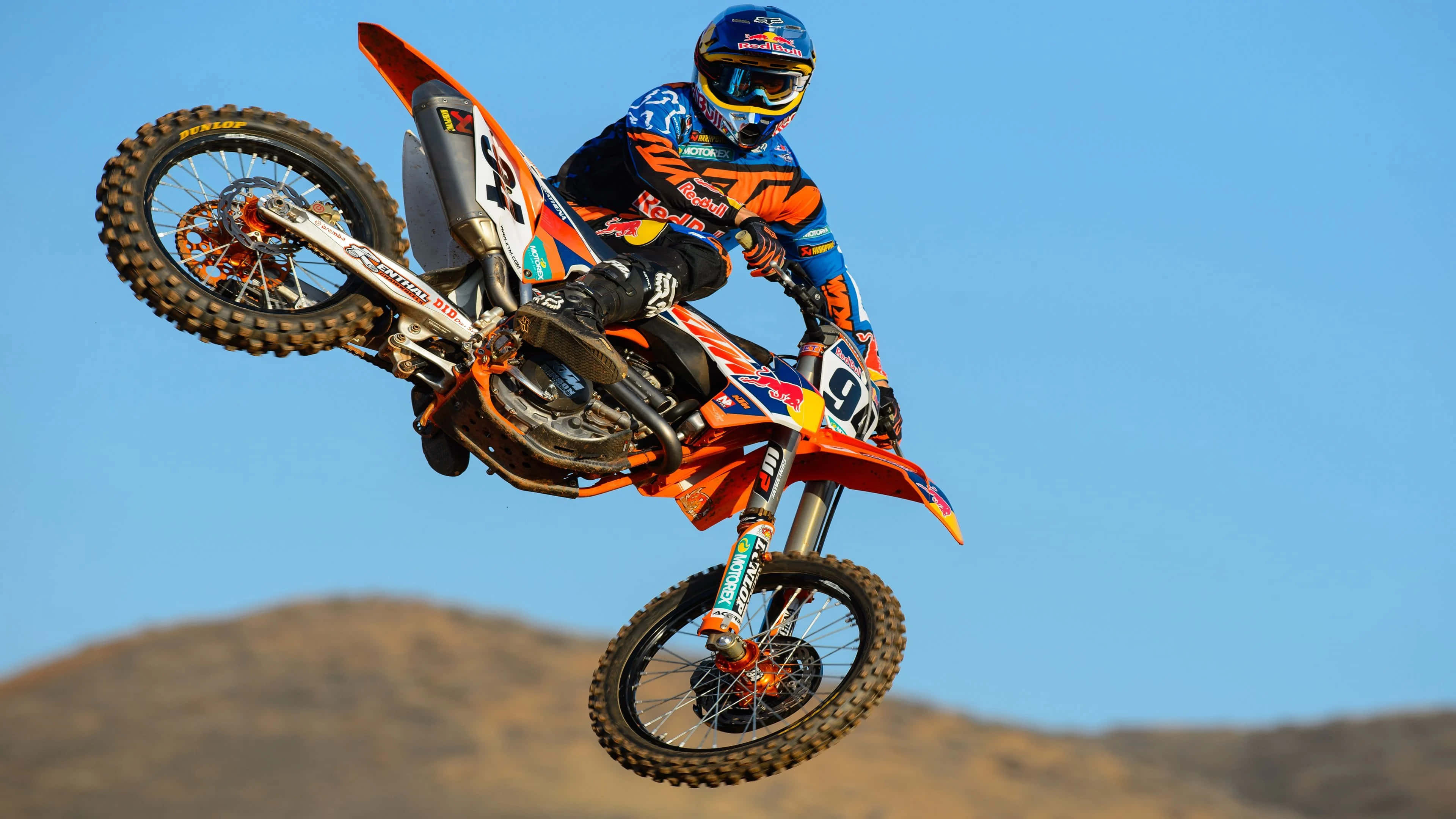Motocross 桌布 壁紙 — 一名身穿鮮豔裝備的越野摩托車手，在藍天白雲和遠山背景下，騎著橙色摩托車在空中飛翔。
