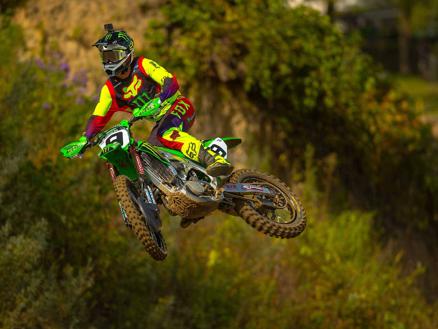 Motocross 桌布 壁紙 — 一名身穿鮮豔裝備的越野摩托車手，騎著綠色摩托車在陽光明媚的森林山坡上空飛翔。