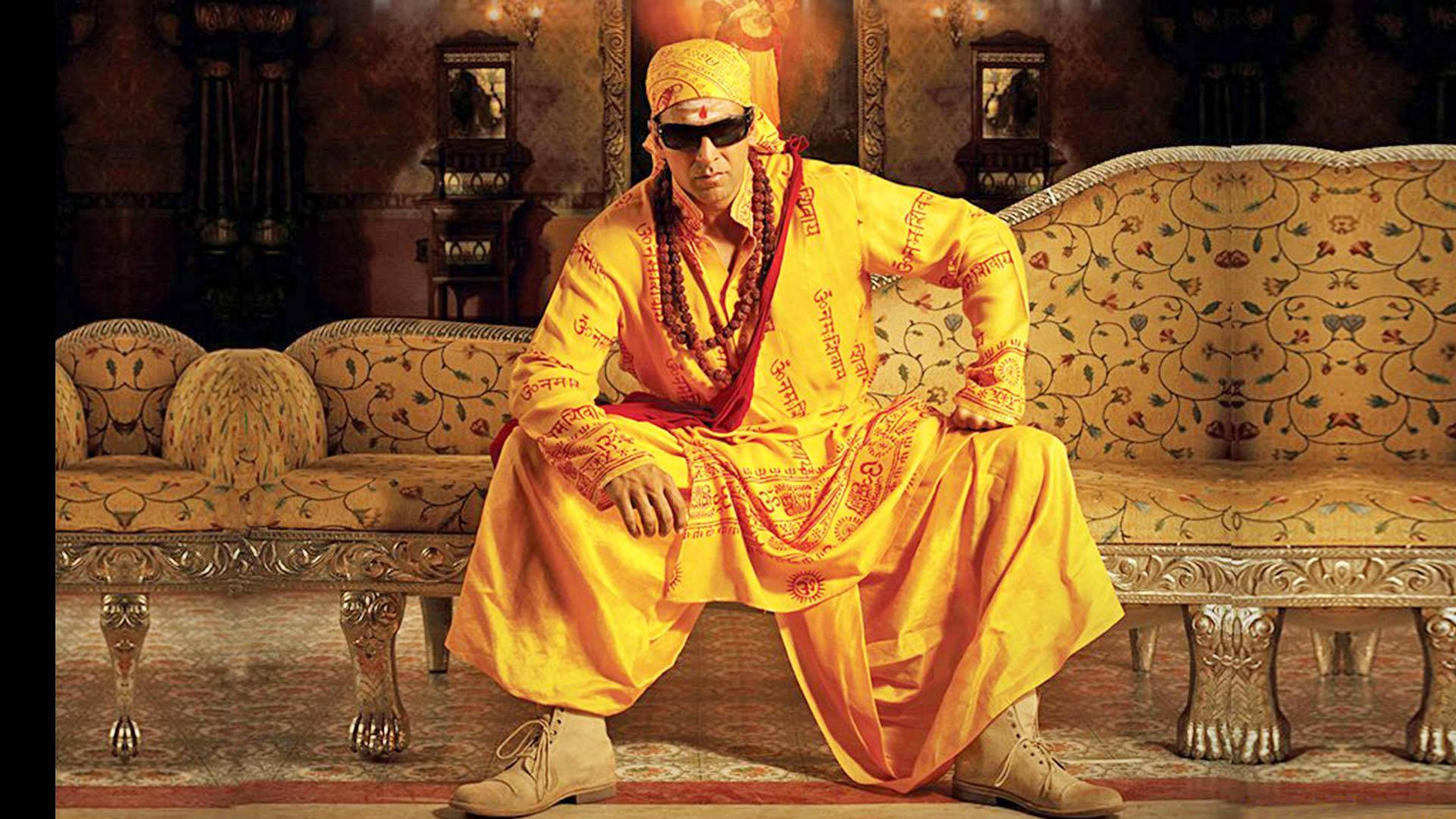 Akshay Kumar 桌布 壁紙 — 一名身穿鮮黃色傳統服飾、戴著太陽眼鏡的男子，自信地坐在一個裝飾華麗的圖案沙發上，身處一個裝飾豐富的房間。