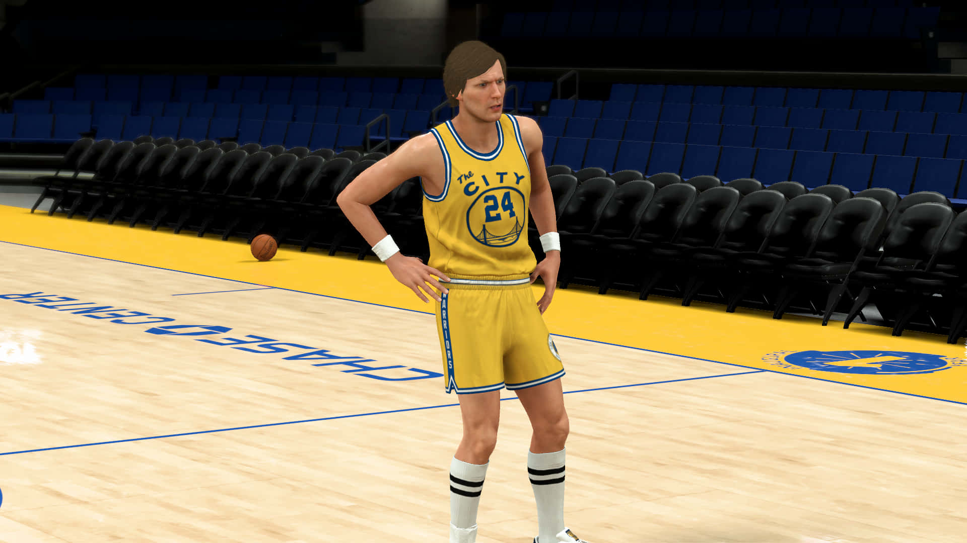 NBA 2k 桌布 壁紙 — 一名身穿黃色'The City'球衣的籃球運動員自信地站在拋光球場上，身後是空無一人的藍色座位。