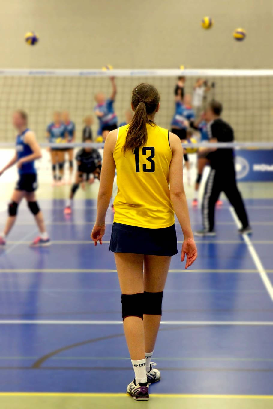 Indoor Volleyball 桌布 壁紙 — 一名身穿黃色13號球衣的女排球員在藍色場地上準備就緒，隊友和對手在她周圍比賽。