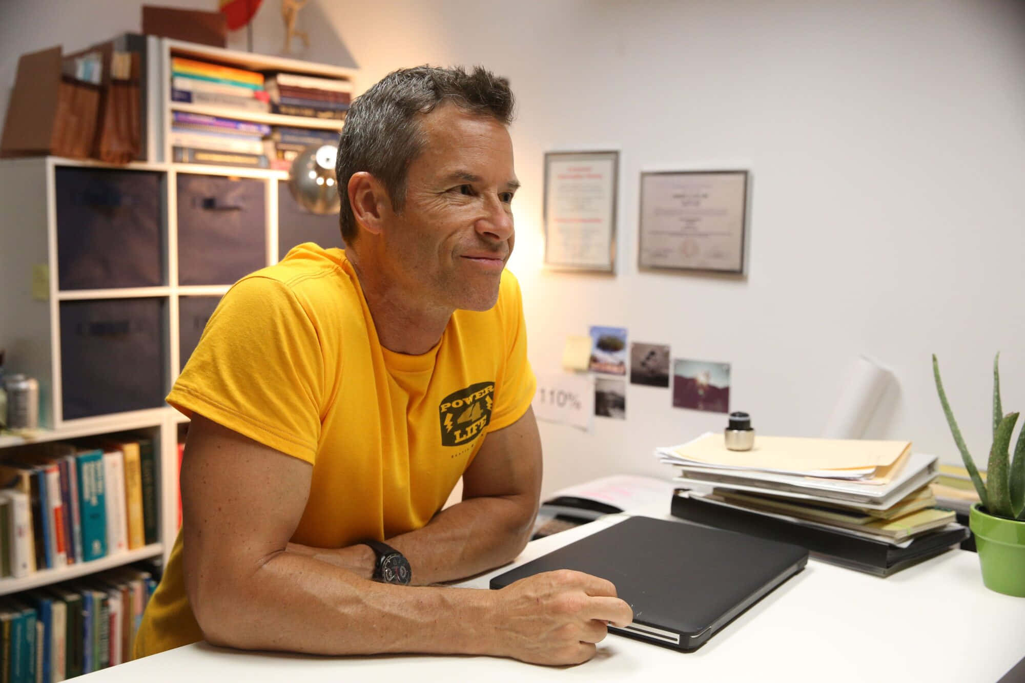 Guy Pearce 桌布 壁紙 — 一名身穿黃色T恤的男子在明亮的辦公室裡，靠著書架和裱框證書的桌子微笑。