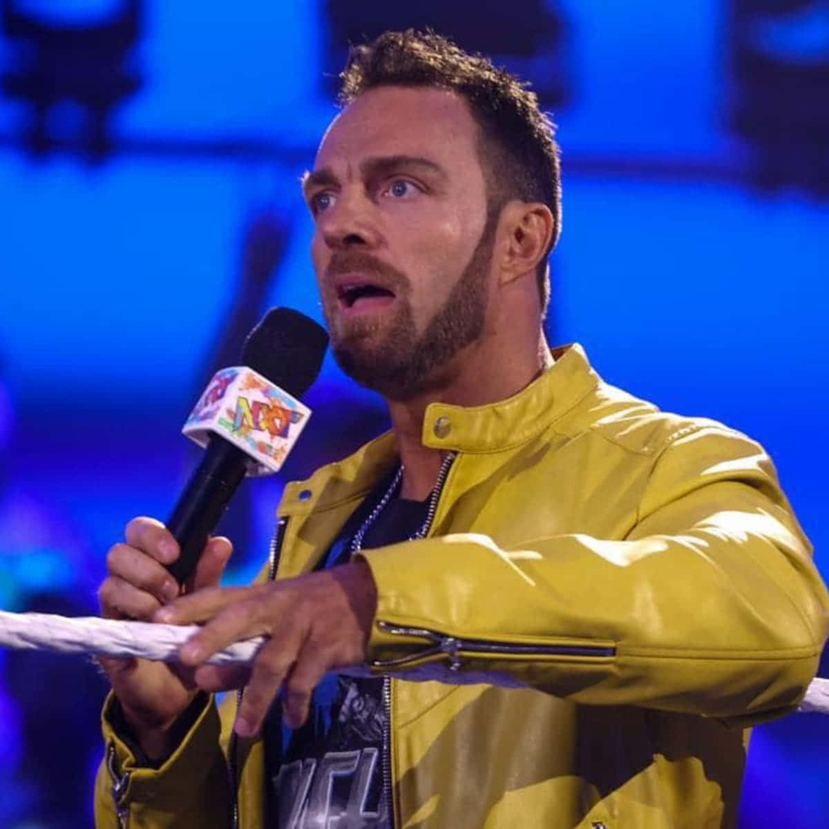 WWE 桌布 壁紙 — 一名身穿黃色外套的摔角手在藍色燈光的擂台上，手握麥克風和繩索，處於戲劇性時刻。