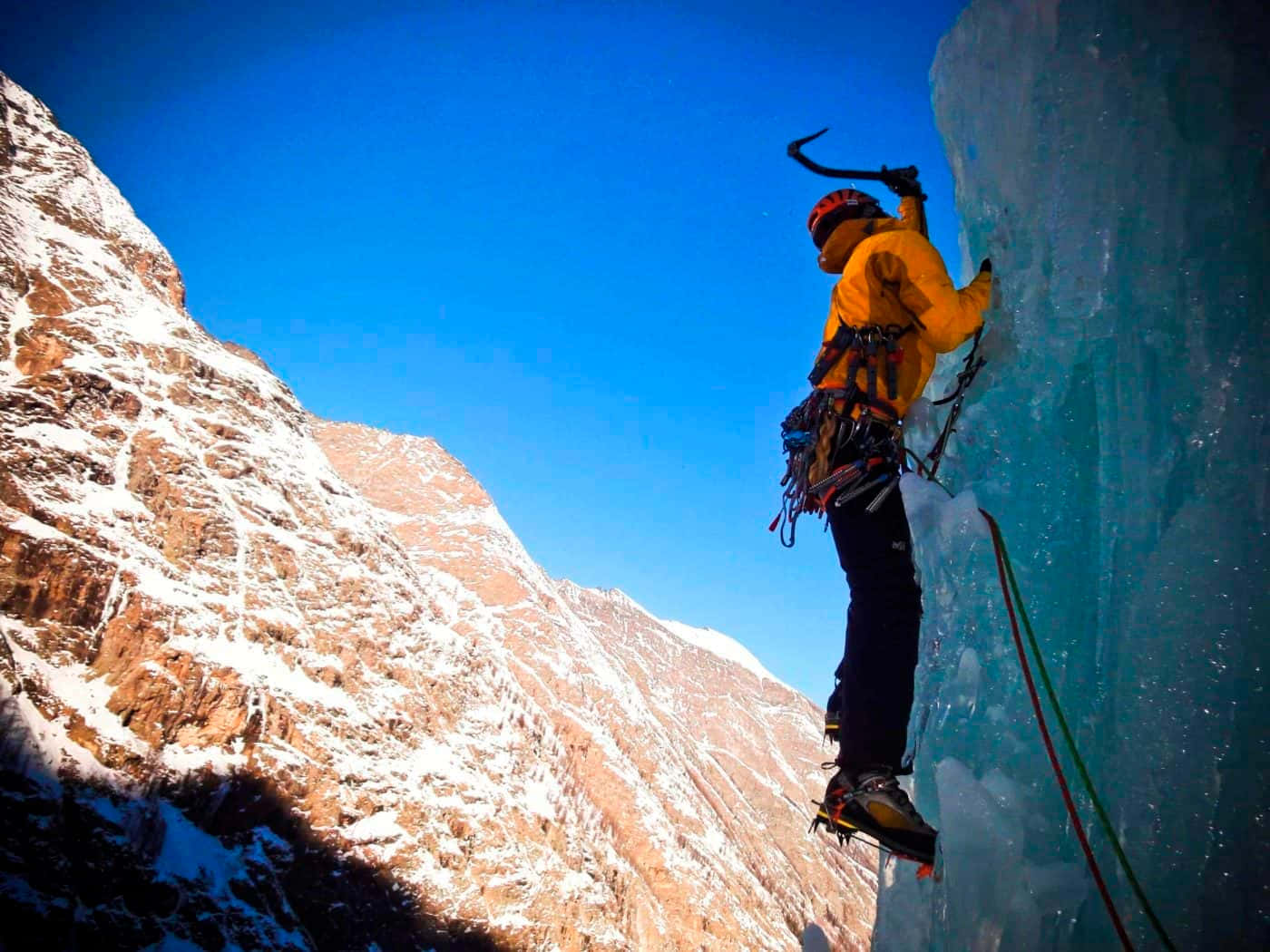 Ice Climbing 桌布 壁紙 — 一名身穿黃色外套的攀岩者，在晴朗的藍天和白雪覆蓋的山脈背景下，攀登高聳的藍色冰壁。