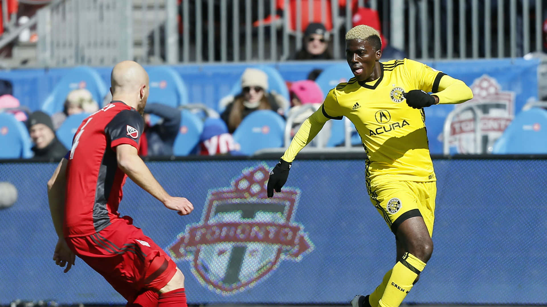 Columbus Crew 桌布 壁紙 — 一名身穿黃色多倫多FC球衣的足球員在陽光明媚的球場上盤球越過身穿紅色的對手。