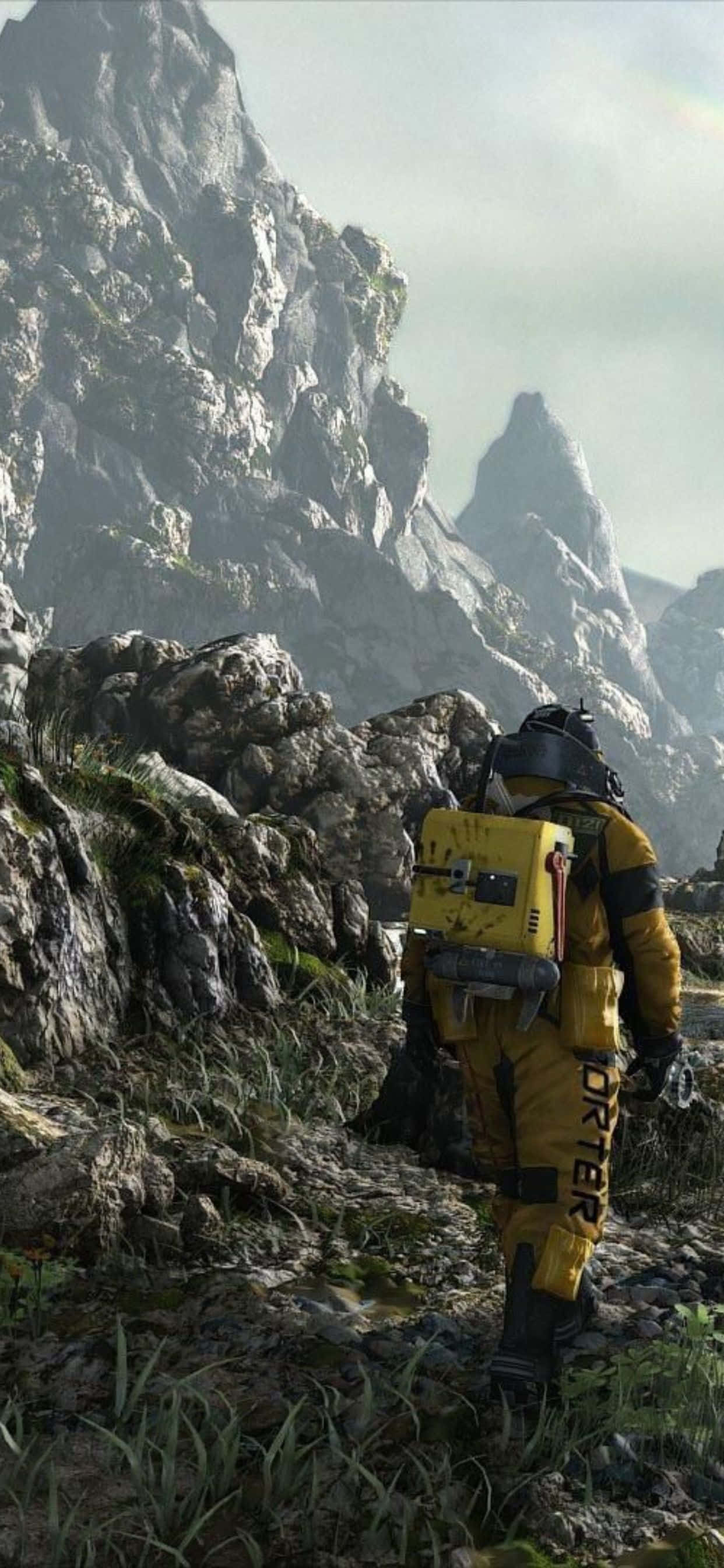 iPhone Xs Max Death Stranding 桌布 壁紙 — 一名身穿黃色套裝的孤獨探險家在朦朧的天空下徒步穿越崎嶇的山地地形。