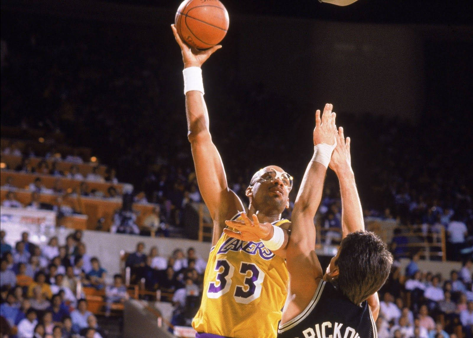 Kareem 桌布 壁紙 — 一名身穿黃色湖人球衣的籃球運動員跳投，而一名身穿黑色球衣的防守球員在明亮的場館燈光下試圖封阻他。