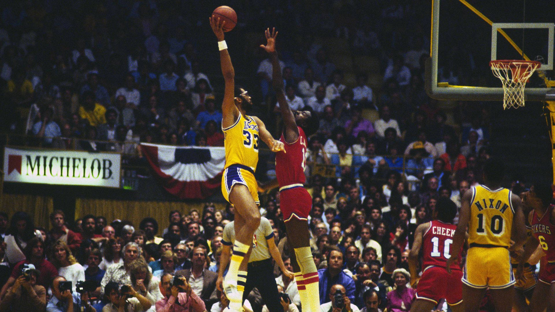 Kareem 桌布 壁紙 — 一名身穿黃色湖人球衣的籃球運動員高高躍起投籃，而一名紅色球衣的防守球員在明亮的場館燈光下試圖封阻他。