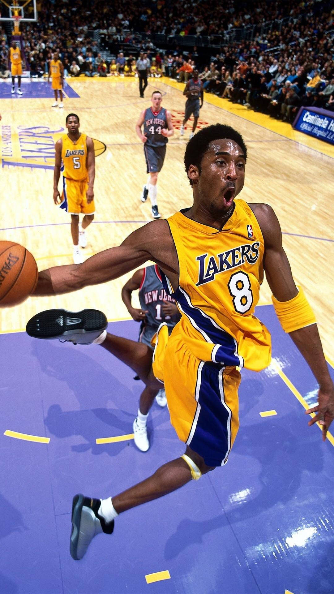 Kobe Bryant iPhone 桌布 壁紙 — 一名身穿黃色湖人球衣的籃球運動員在明亮的場館燈光下，於激烈的比賽中持球躍起空中。