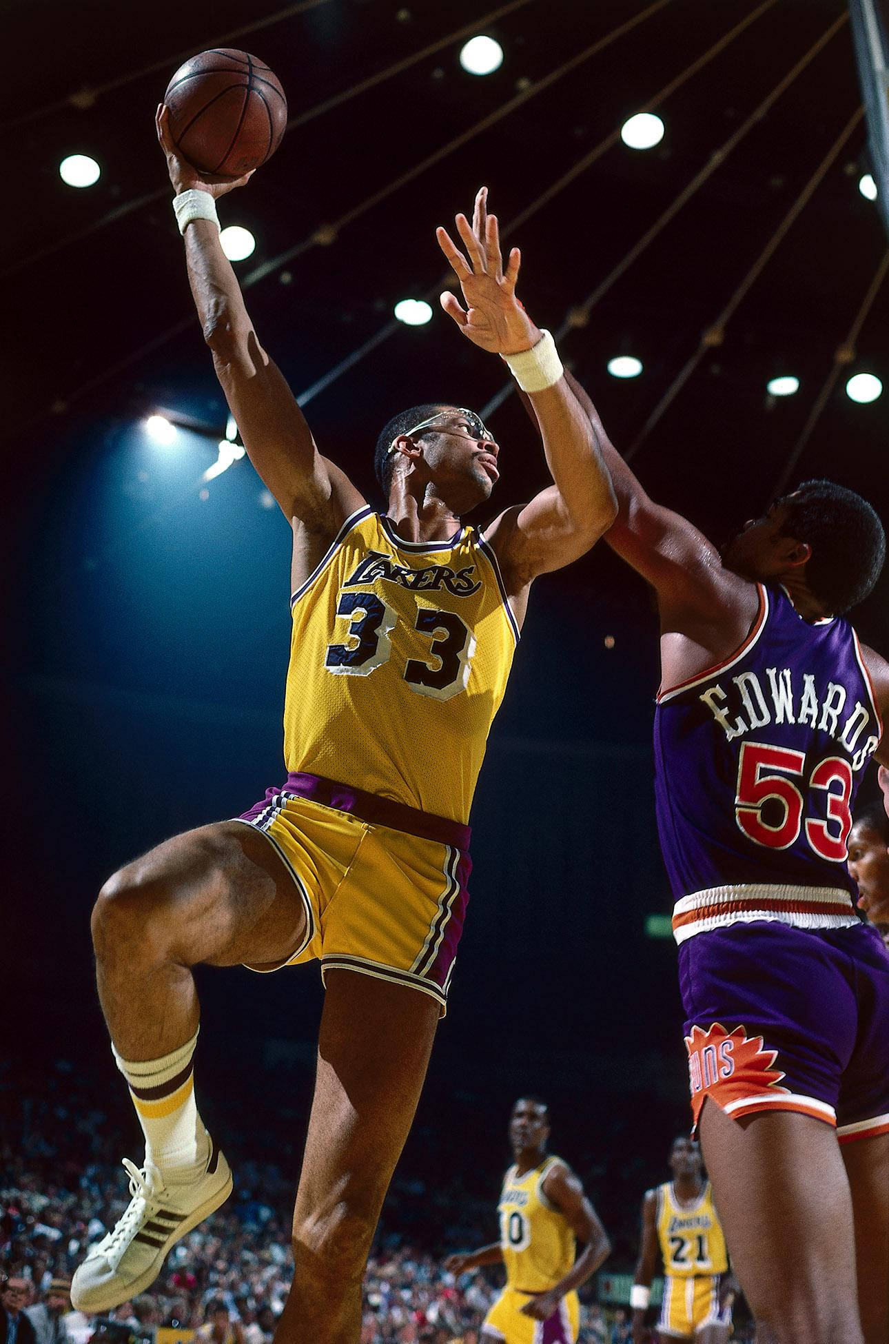 Kareem 桌布 壁紙 — 一名身穿黃色湖人球衣的籃球運動員高高躍起投籃，而一名身穿紫色球衣的防守球員試圖封阻。