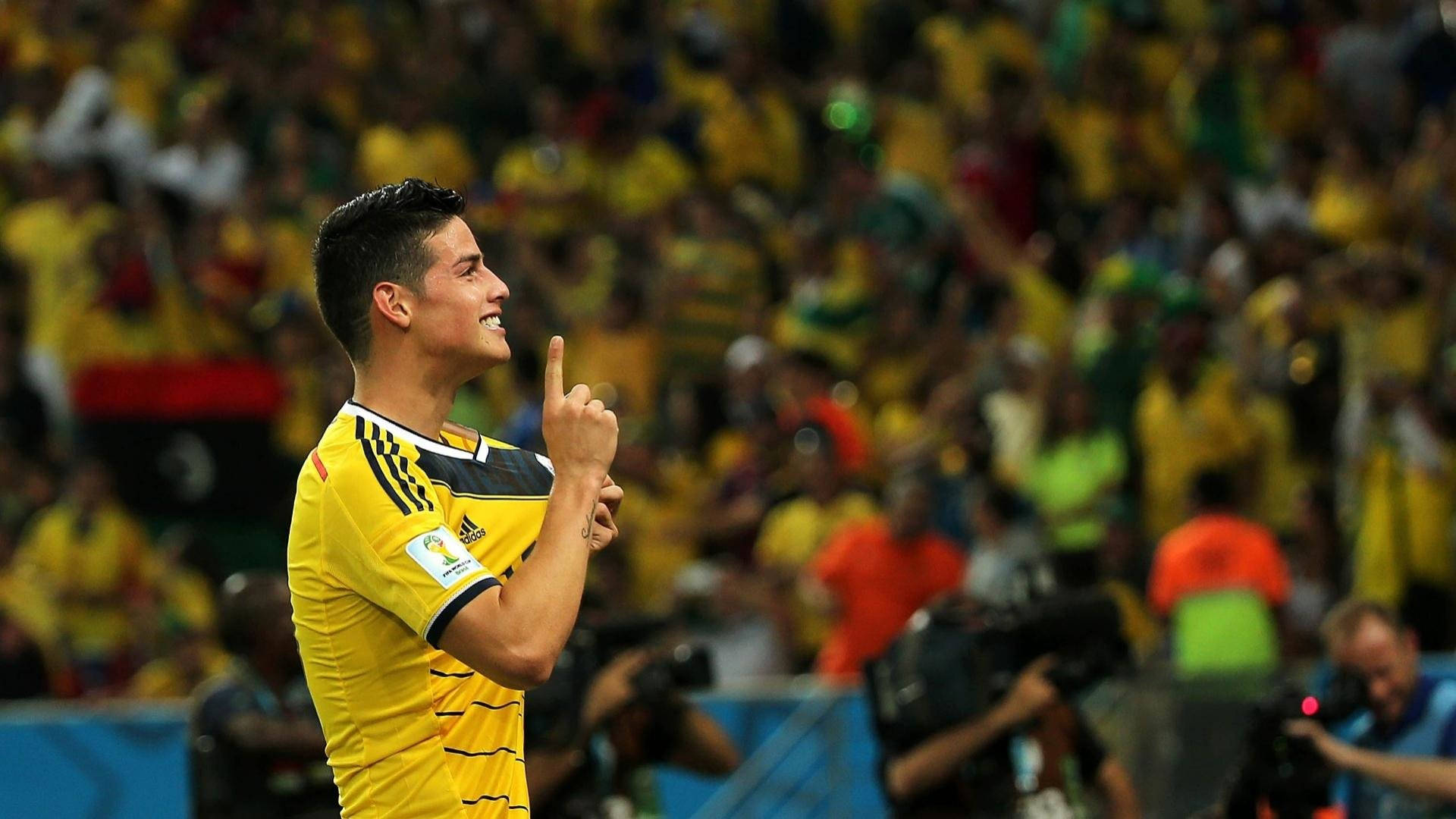 James Rodriguez 桌布 壁紙 — 一名身穿黃色球衣的哥倫比亞足球員豎起手指慶祝，身後球場內的球迷歡呼雀躍。