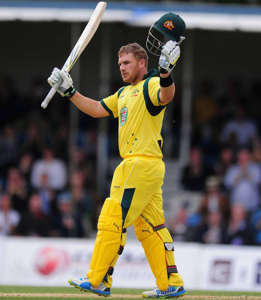 Aaron Finch 桌布 壁紙 — 一名身穿黃色球衣的板球運動員在陽光明媚的球場上，背景是模糊的觀眾，他高舉球棒和頭盔，慶祝勝利。